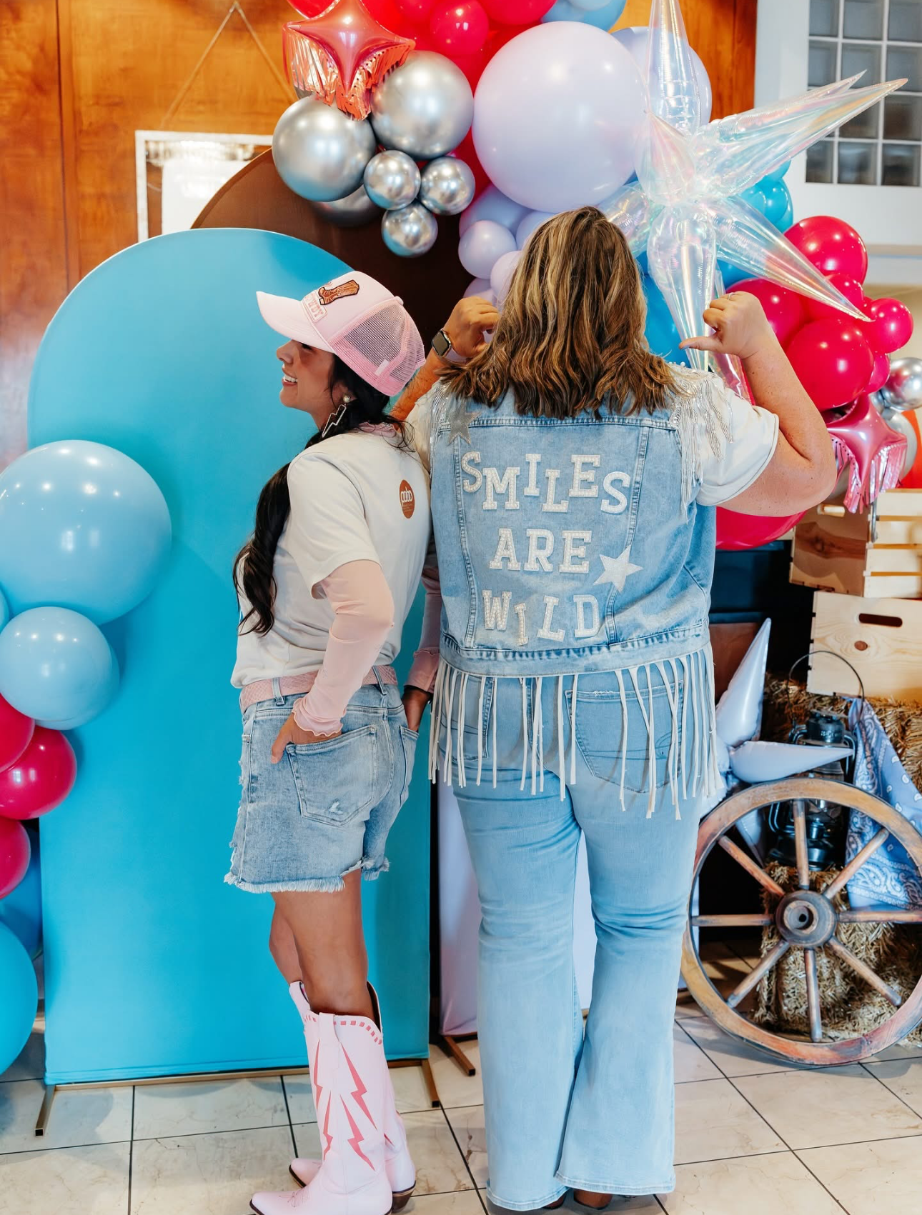 Two people pose near a balloon arch. One wears a denim vest with 