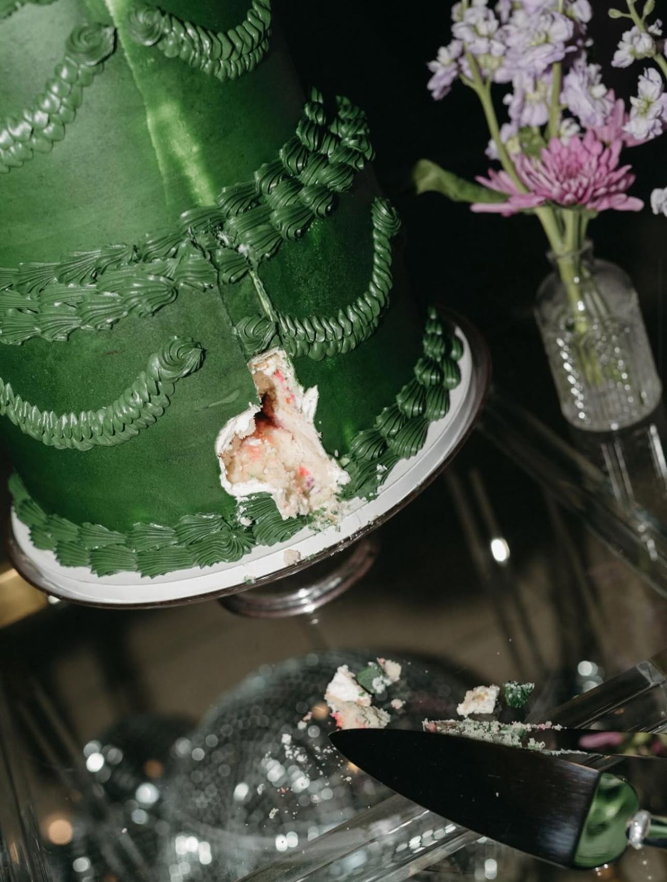 Green decorated cake with missing piece, on a glass table with flowers. Cake server nearby.