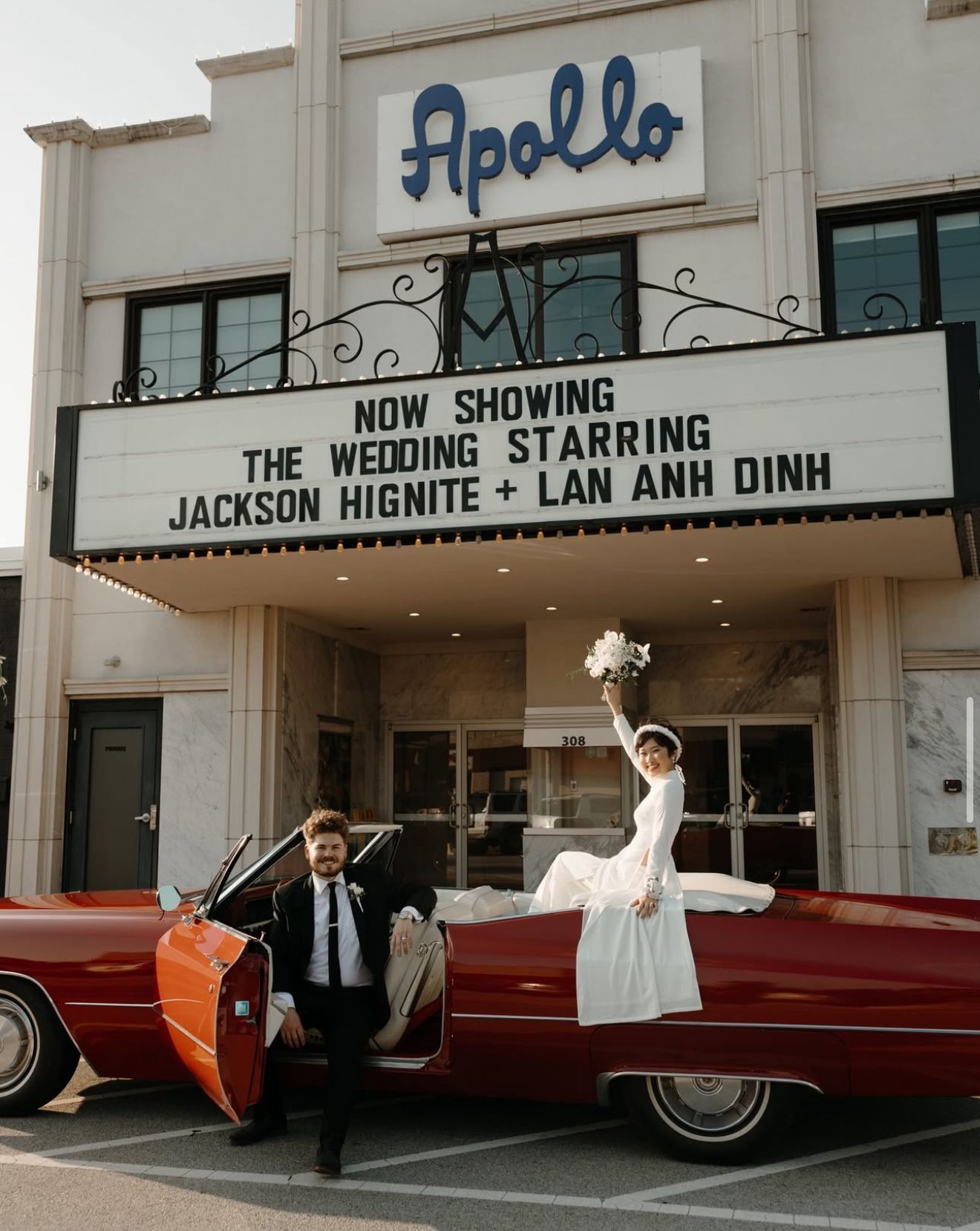 Couple in wedding attire posing by a red vintage car in front of the Apollo theater marquee.