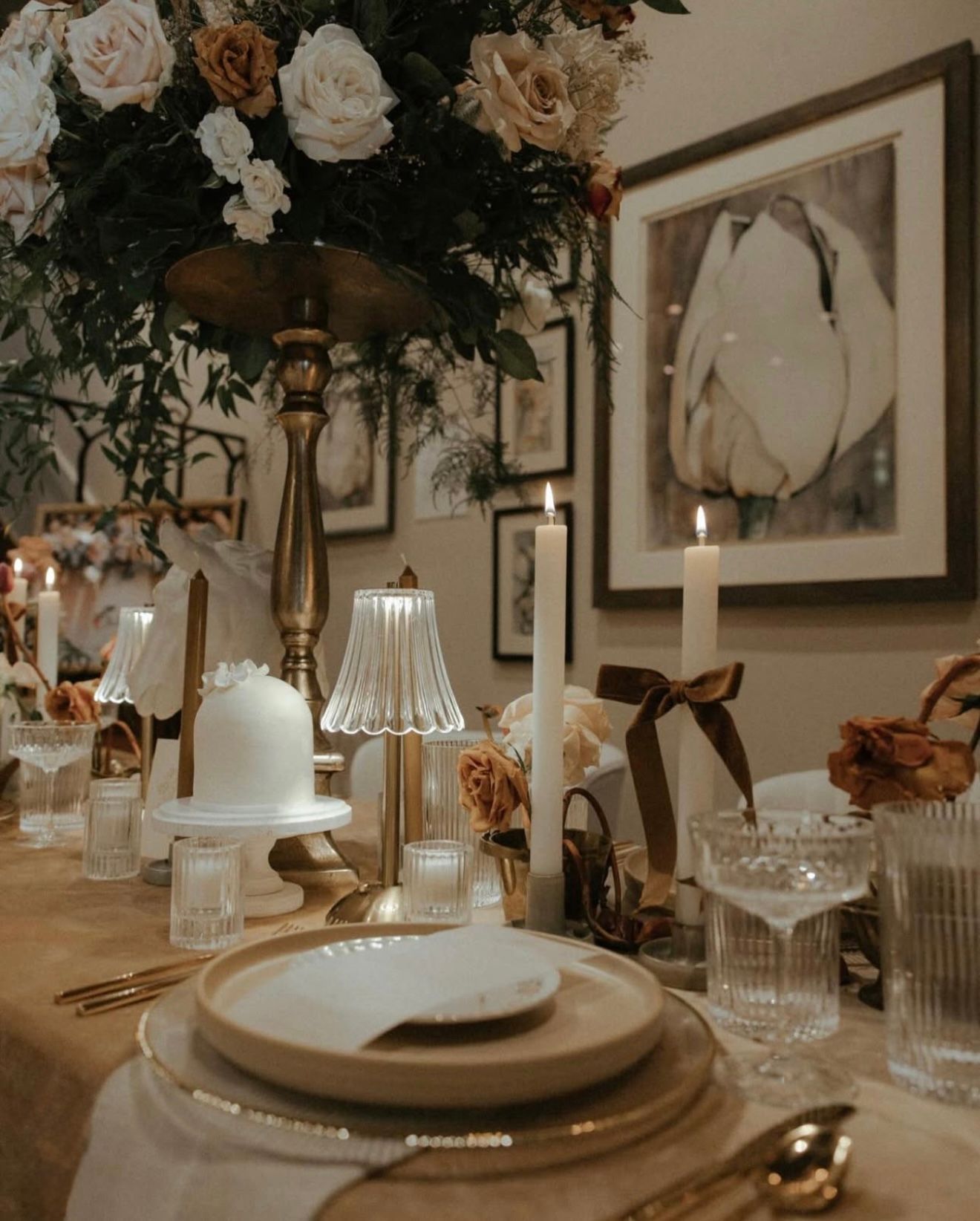 Elegant dining table set for a special occasion.  Floral centerpiece, candles, framed artwork, and place settings with gold accents.