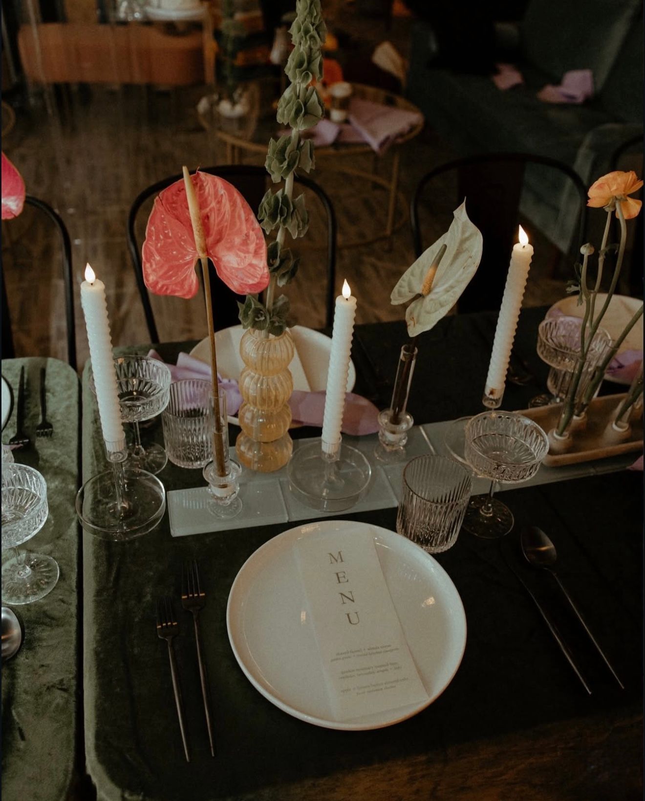 Elegant table setting with candles, flowers, and a menu on a white plate; dark green tablecloth.