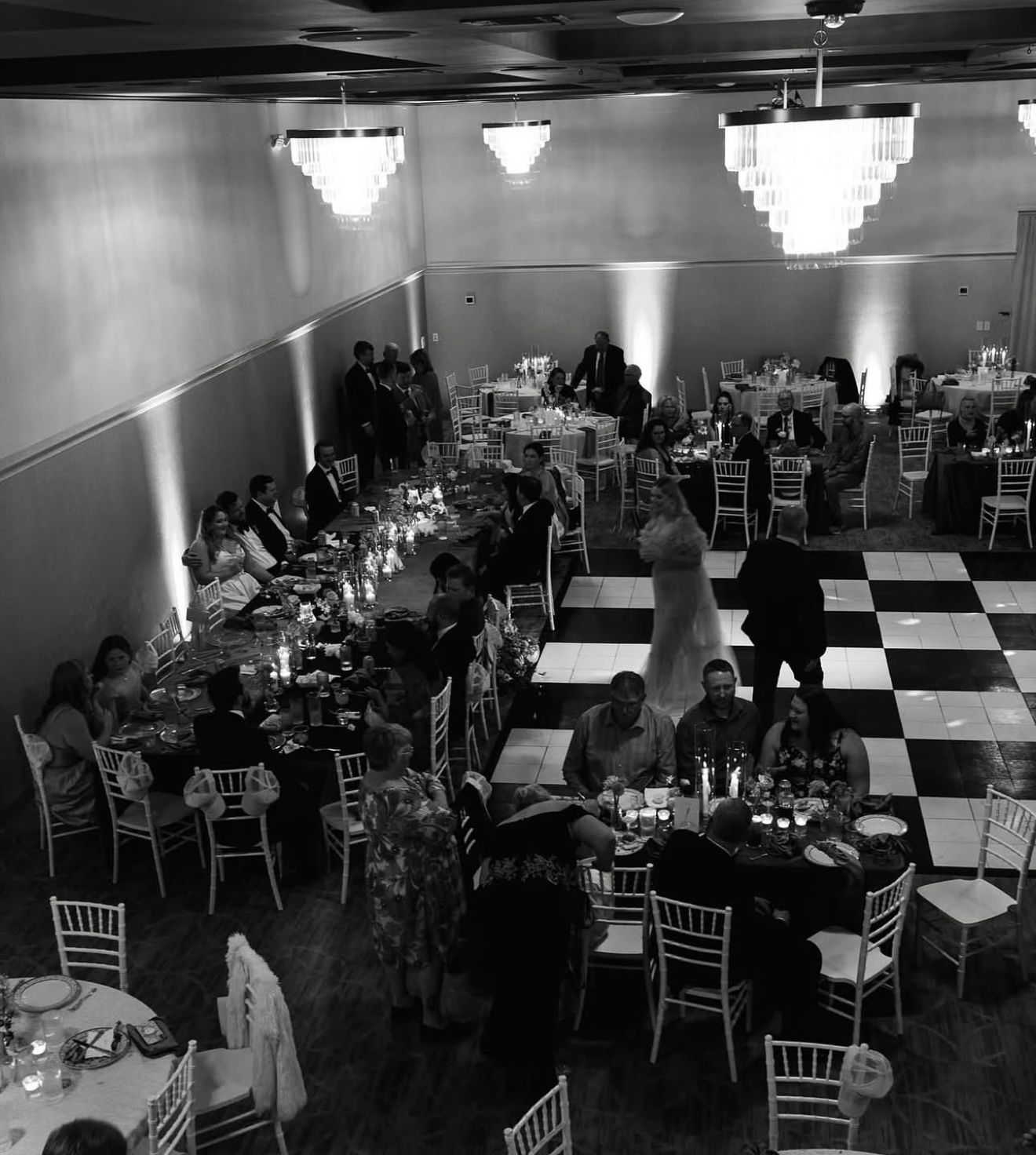 Black and white photo of a wedding reception in a large hall with tables set for guests. A couple dances on a checkered dance floor.
