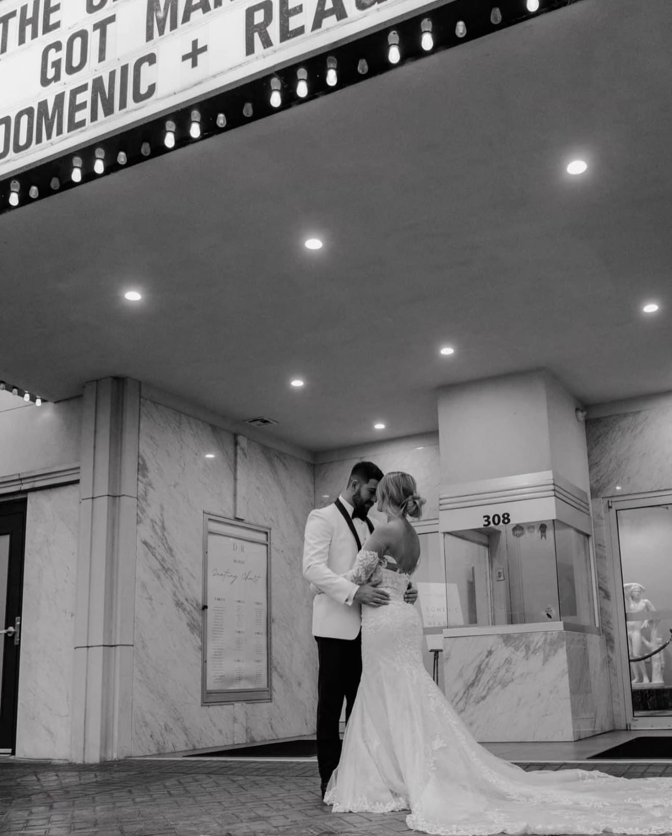Newlyweds embrace in front of a theater. Woman in a wedding dress, man in a tuxedo. Black and white.