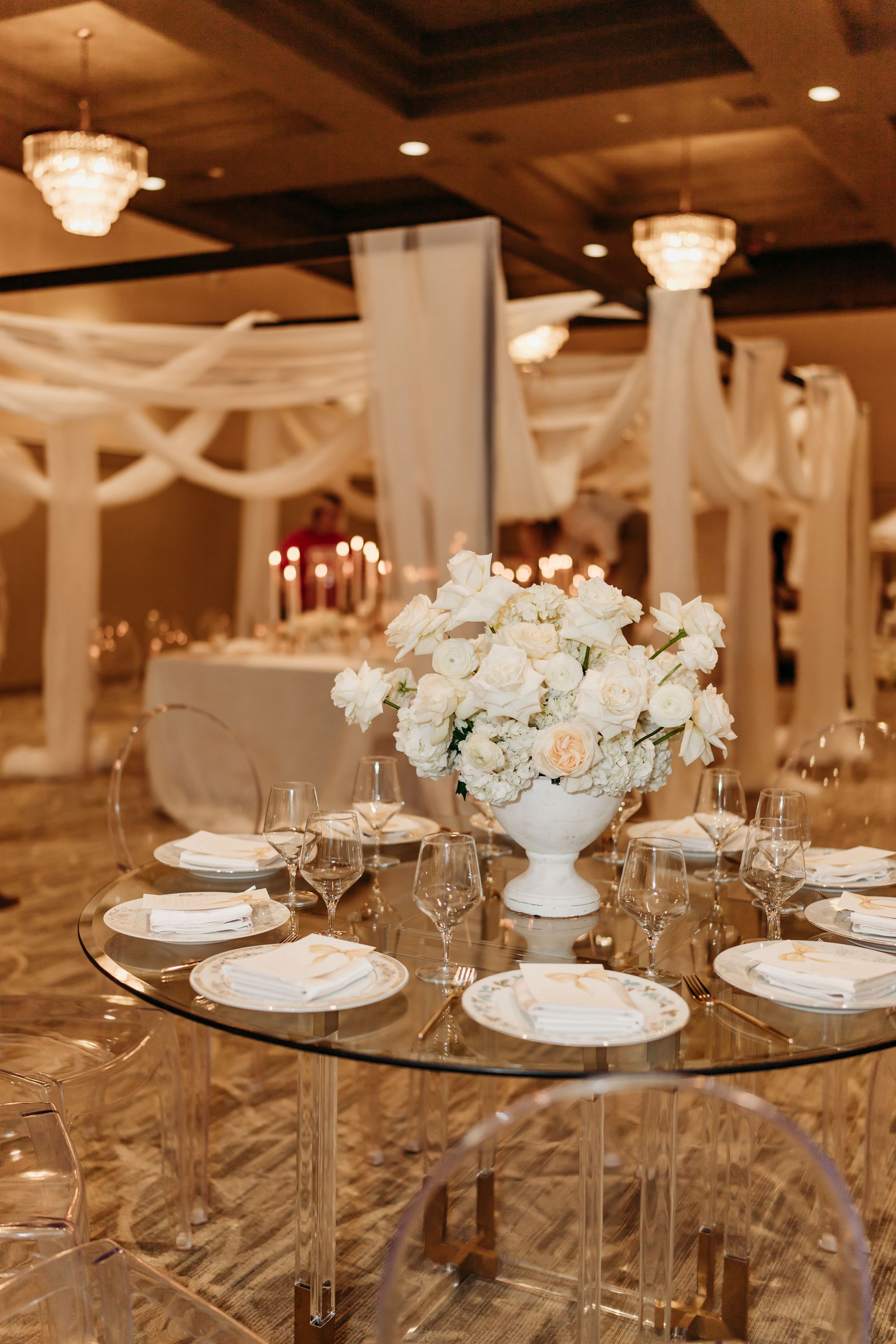 Elegant wedding reception table with white floral centerpiece and clear chairs.