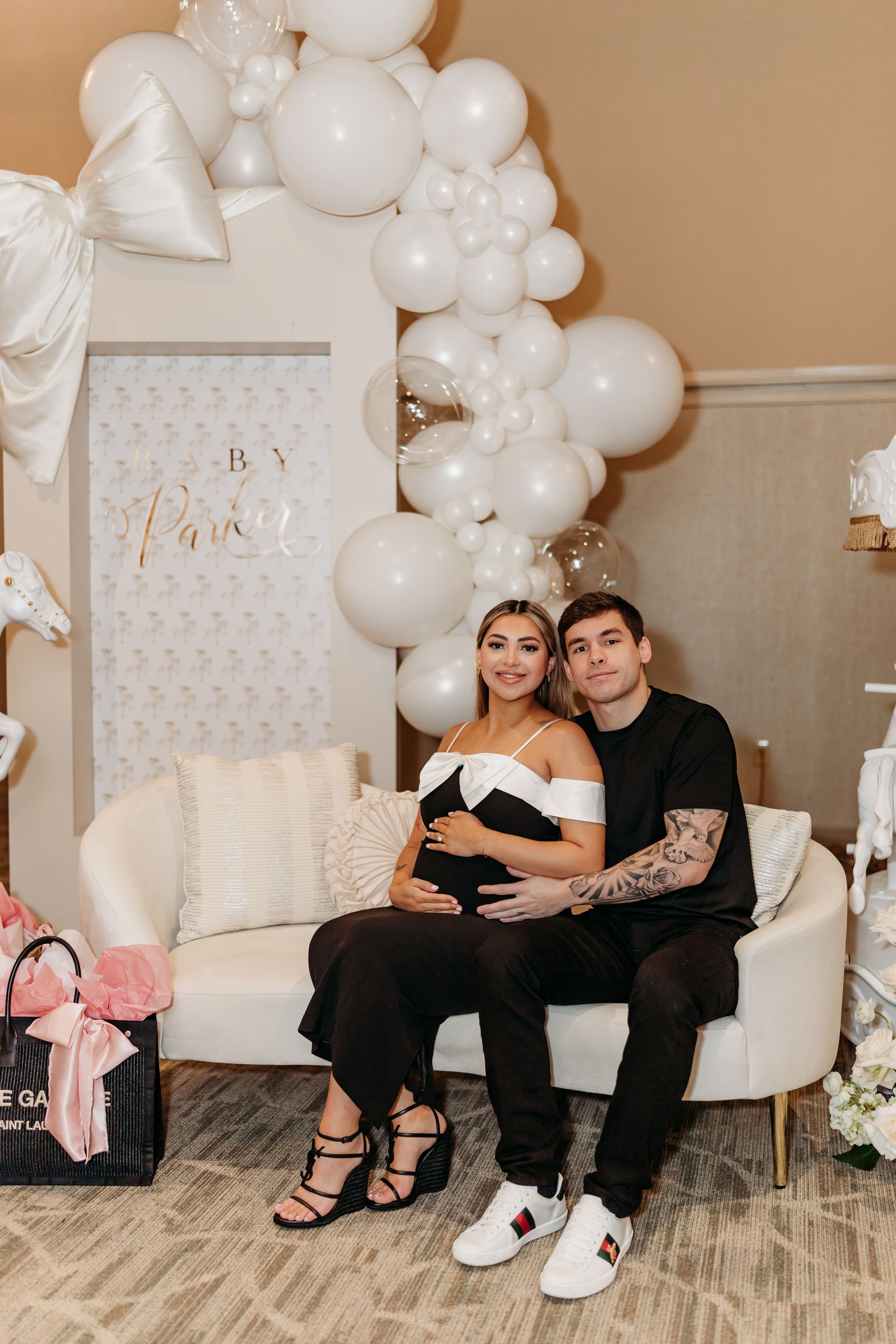 Pregnant woman and partner seated on a couch, surrounded by balloons and baby shower decor.