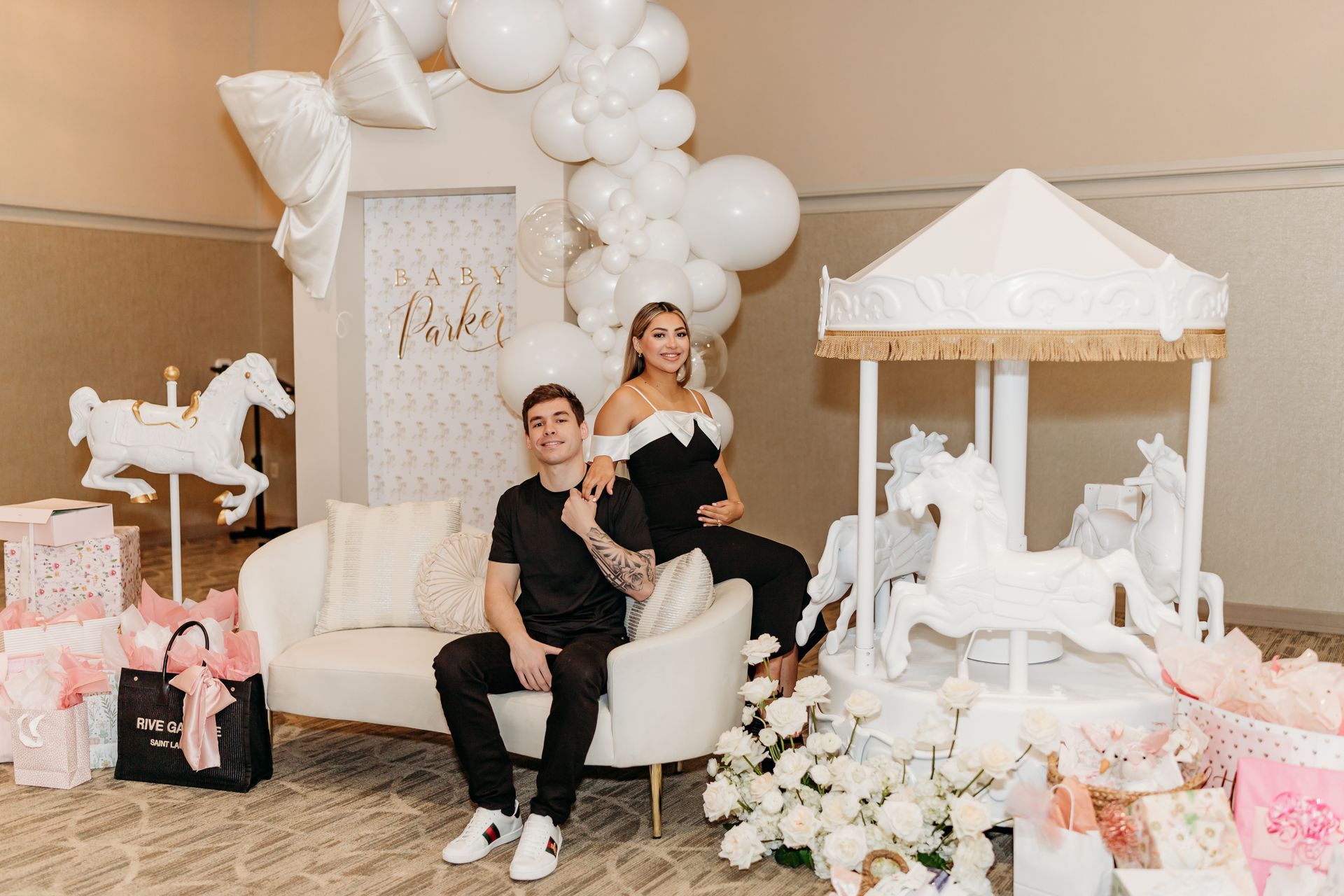 Women in a wood-paneled room, conversing and holding drinks near a floral arrangement, with a window and bar area in view.