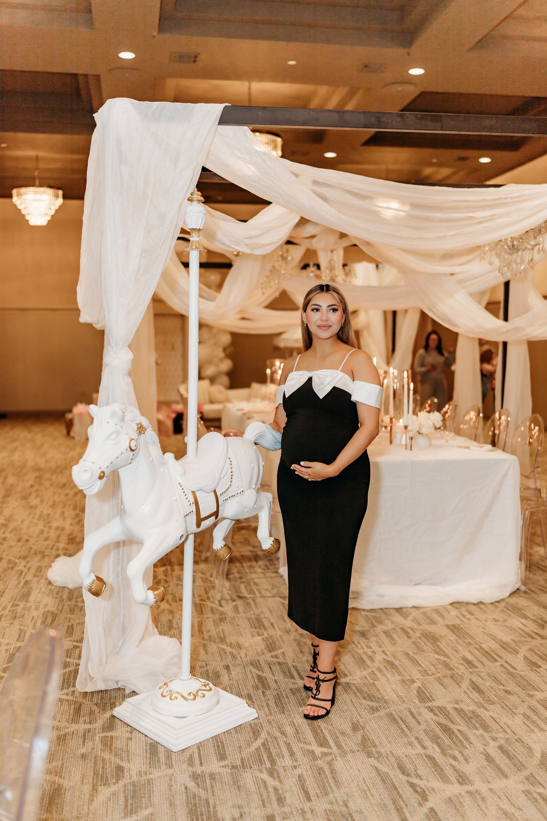 Pregnant woman in black dress at a party, standing by a carousel horse decoration.