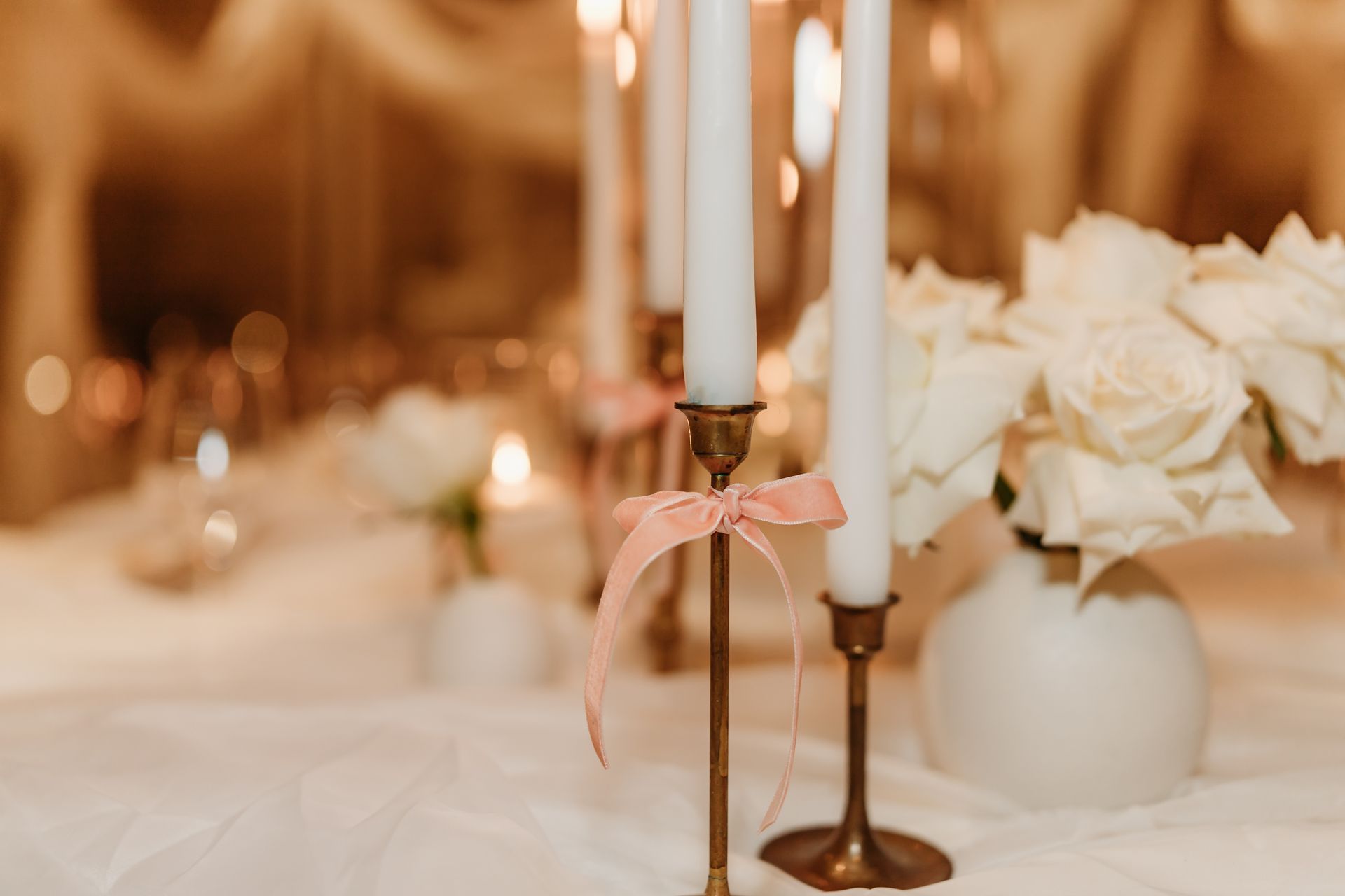 Elegant table setting: white candles, pink ribbons, white roses, and gold candle holders.