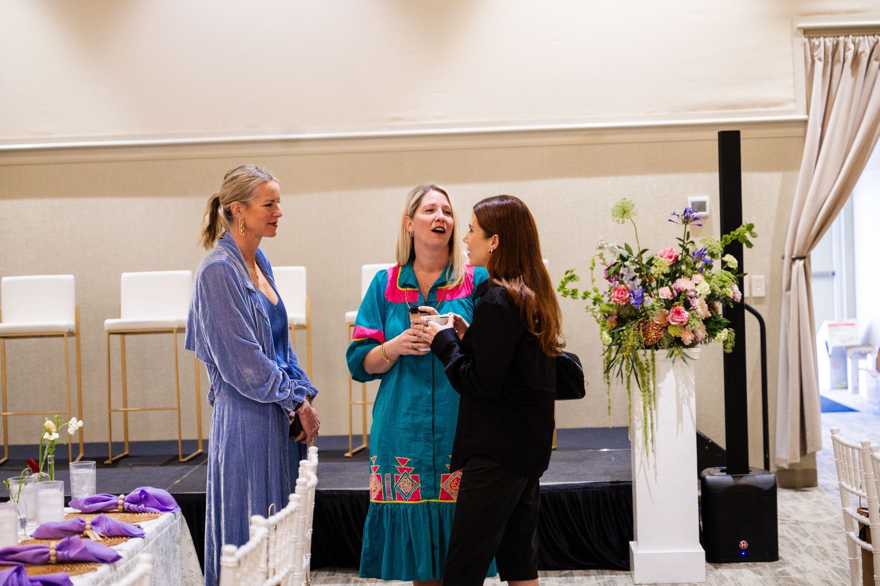 Three women chatting near a stage with flowers and tables set for a meal.