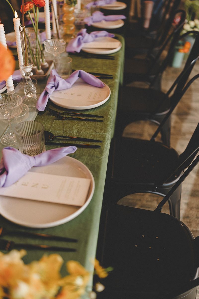 Long table set for a meal with plates, napkins, and black chairs. Green tablecloth, flowers, and candles.