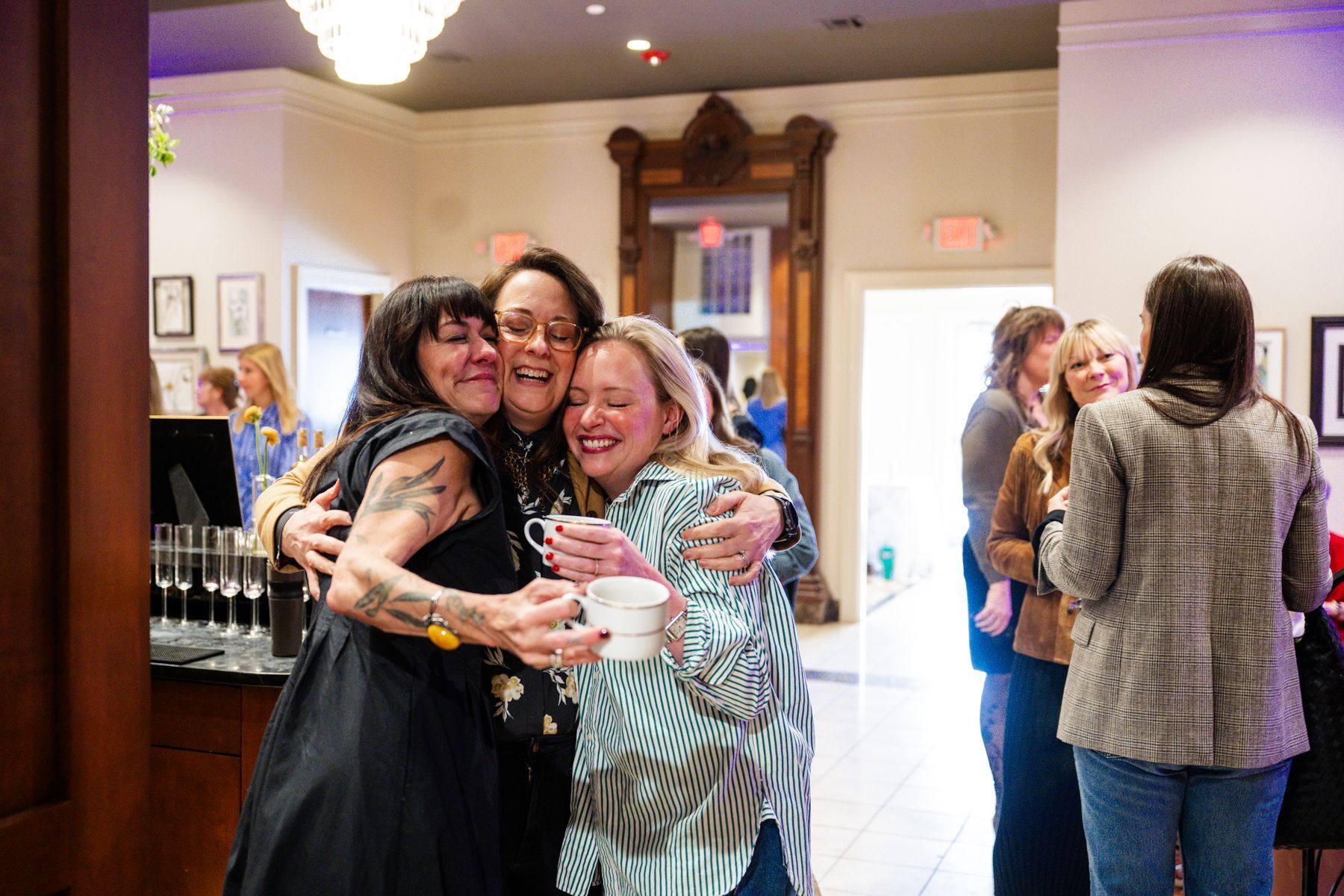 Three women embrace, laughing, holding mugs, indoors with other people.