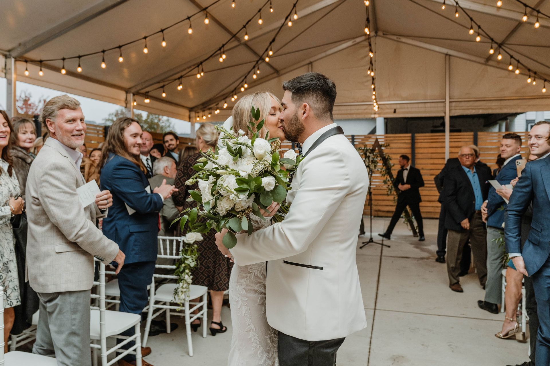 Bride and groom kissing under tent after wedding ceremony, surrounded by guests.