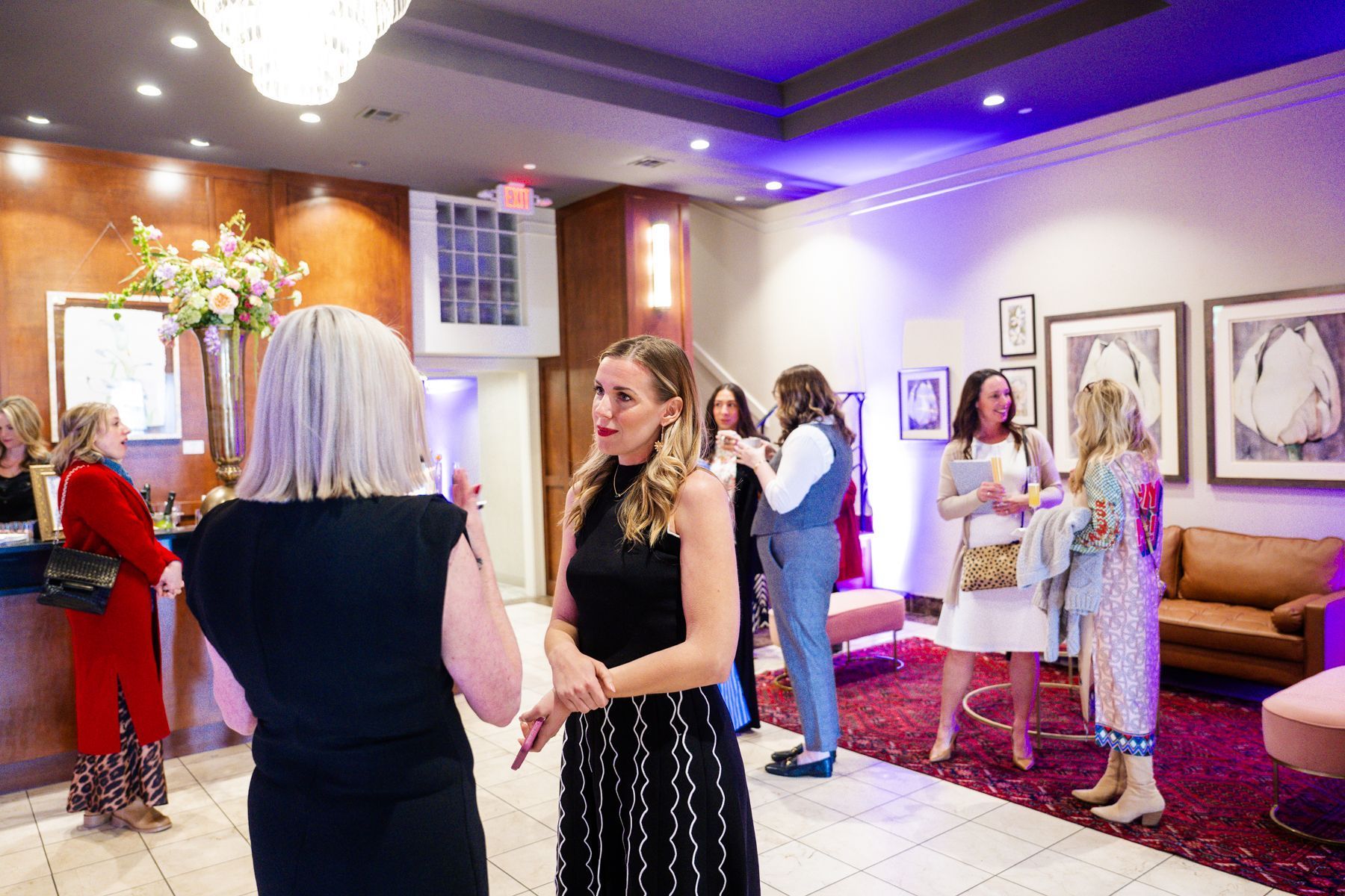 People in an elegant lobby chatting at an event. Gold and white decor, artwork on walls, red rug, and dim lighting.