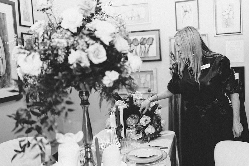 Woman adjusting large floral arrangement on a decorated table. Artwork hangs on the wall in the background.