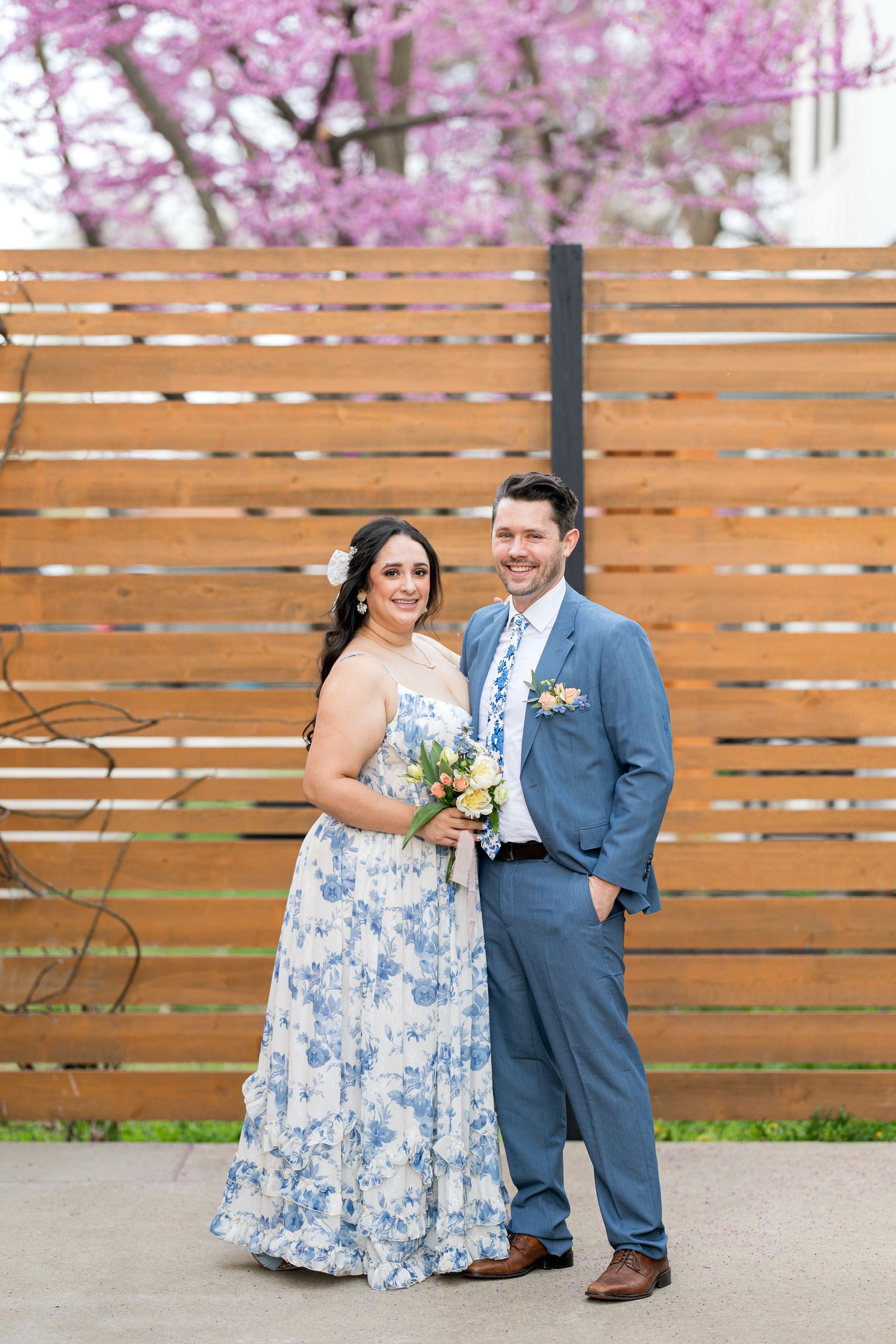 Couple poses in front of wooden fence, woman in blue floral dress, man in blue suit, smiling.