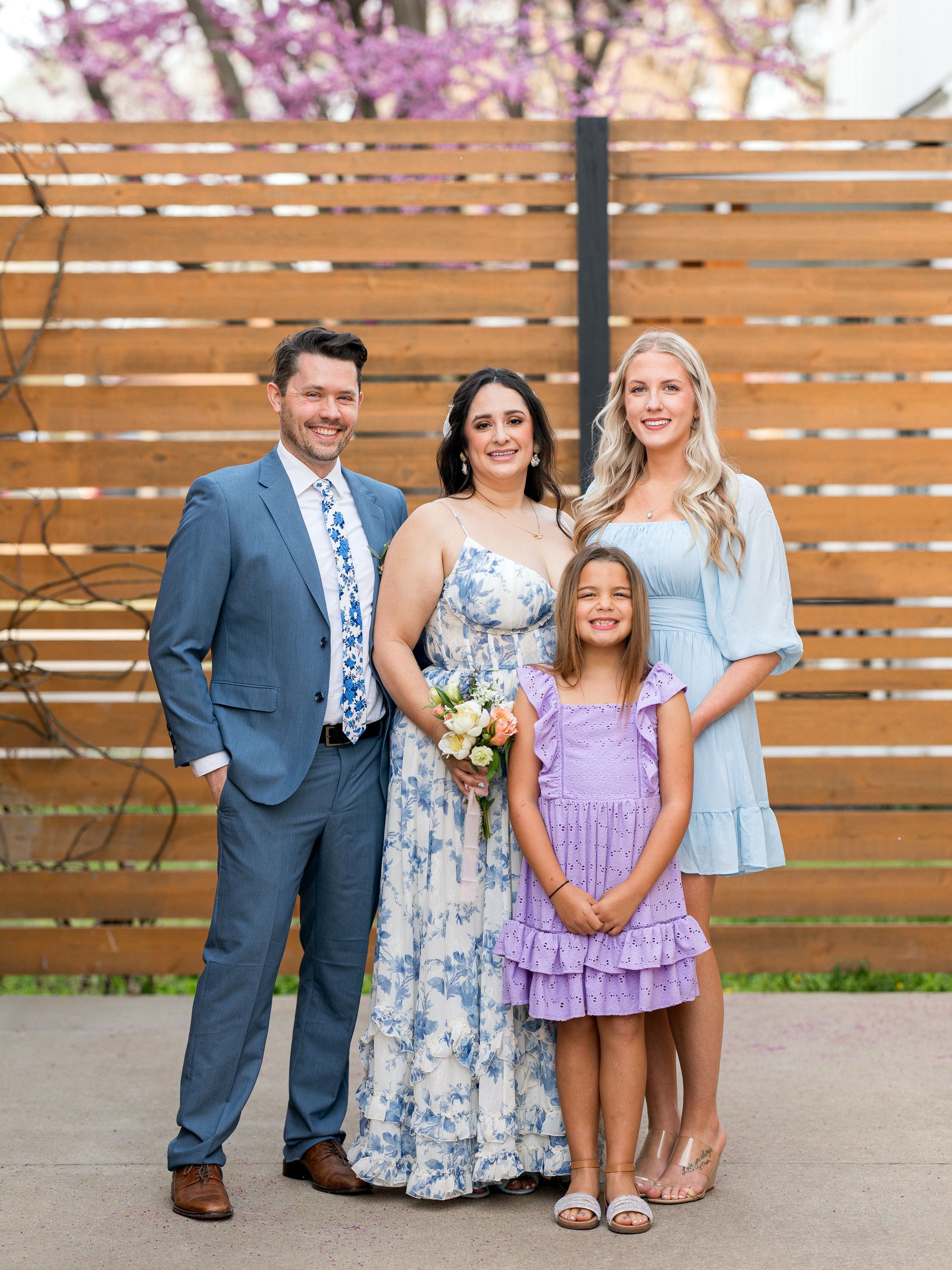 Family of four smiling, posing for a photo. Woman in blue floral dress holds bouquet, others in blue outfits, standing outside.