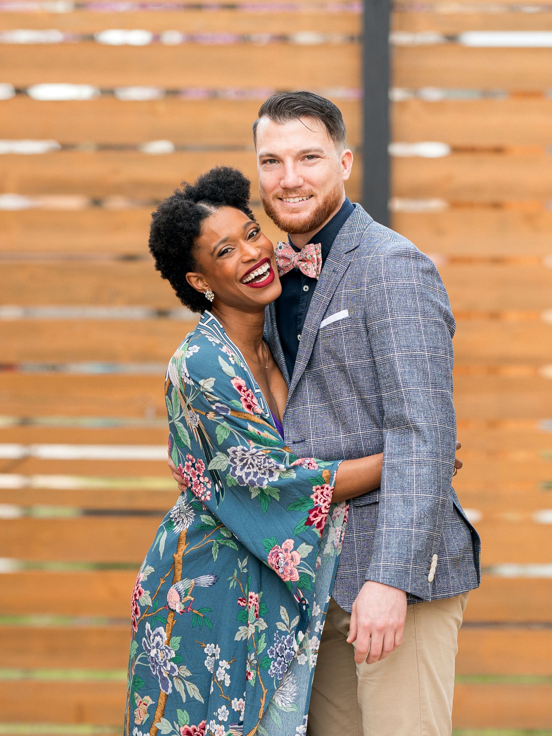 Couple smiling, embracing. Woman in floral robe, man in patterned jacket, wood backdrop.
