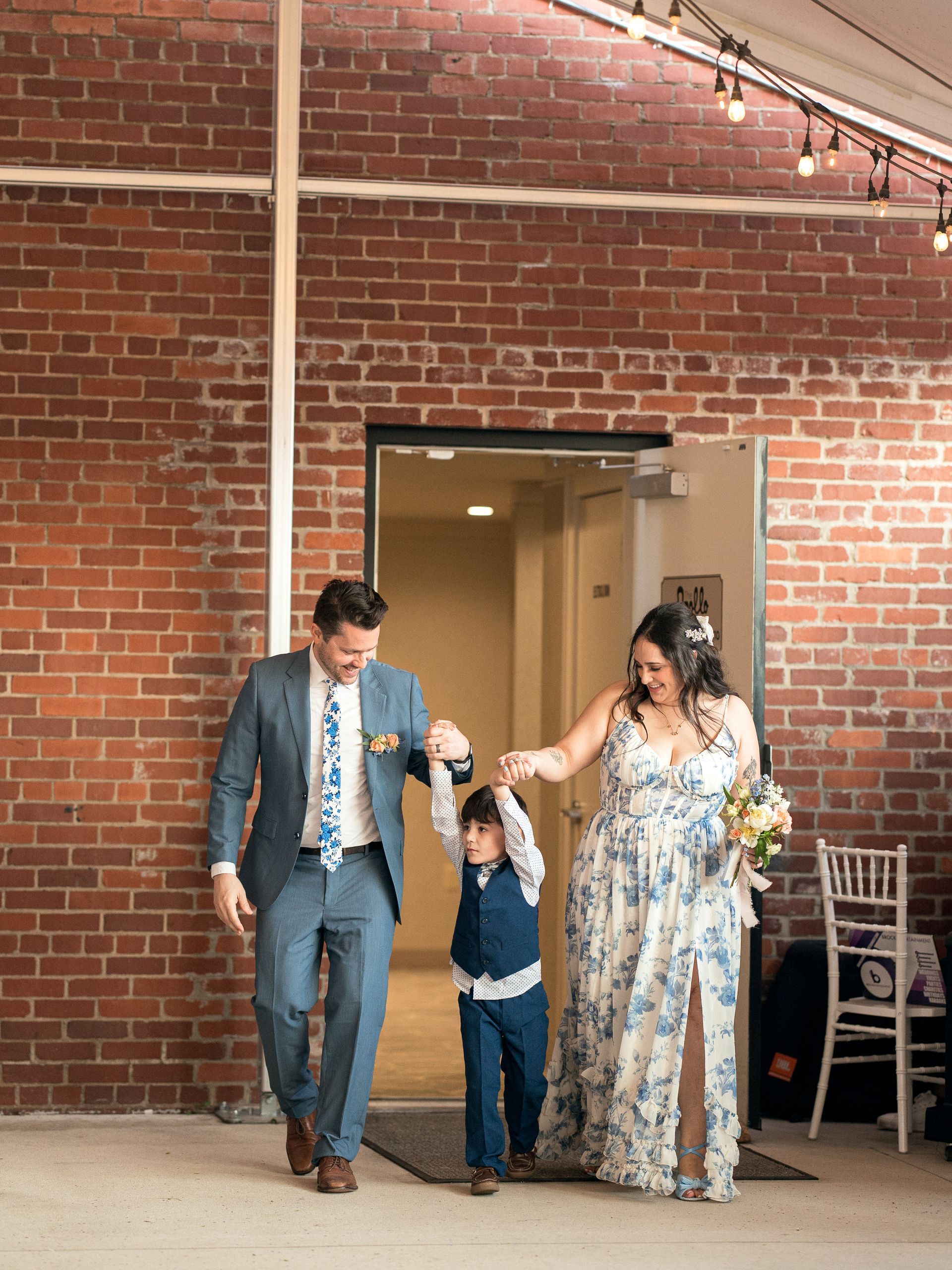 Newlyweds and child exit a door, smiling and holding hands, brick wall in background.