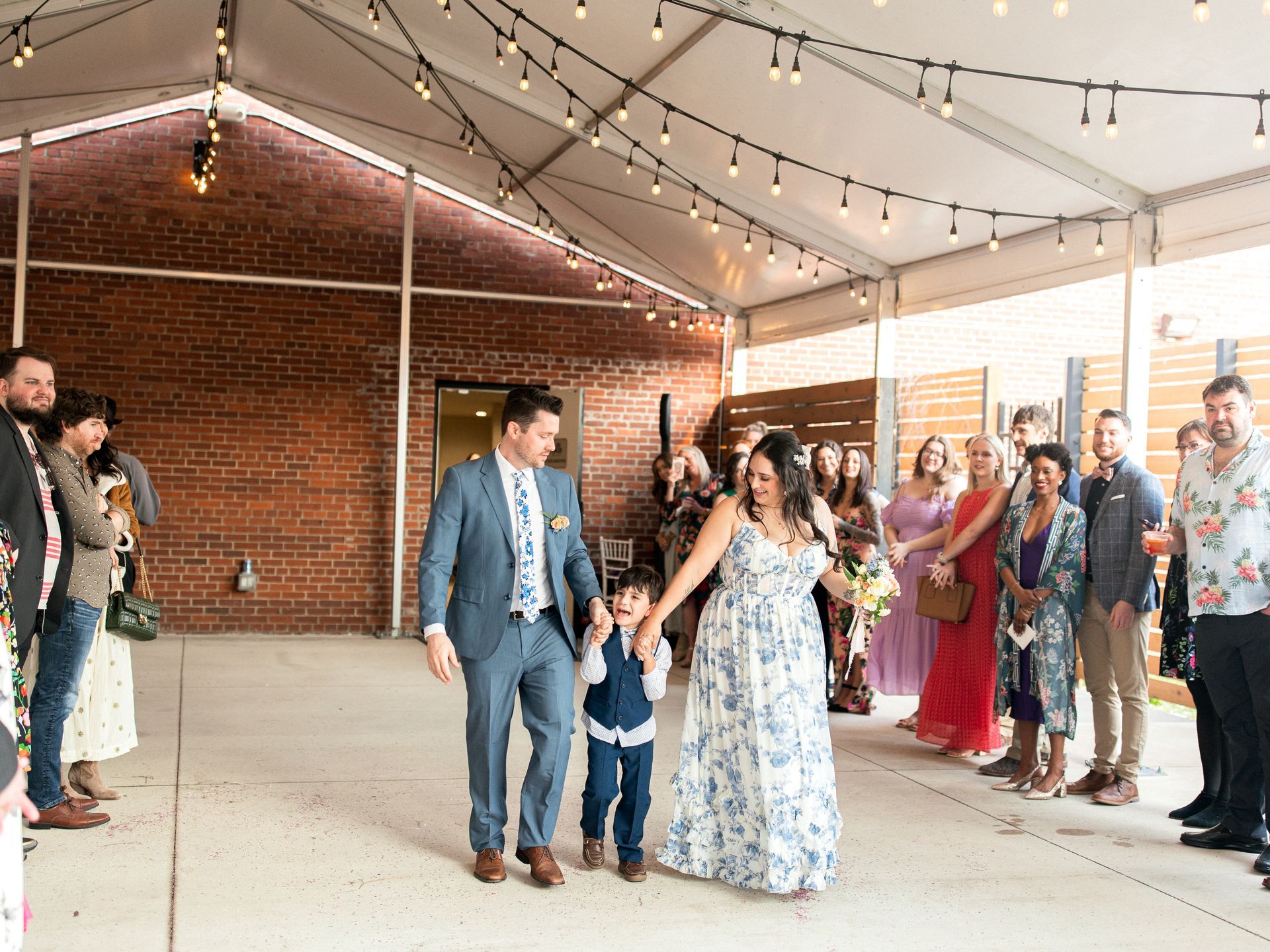 Bride, groom, and child walk down aisle at outdoor wedding reception; guests cheer.