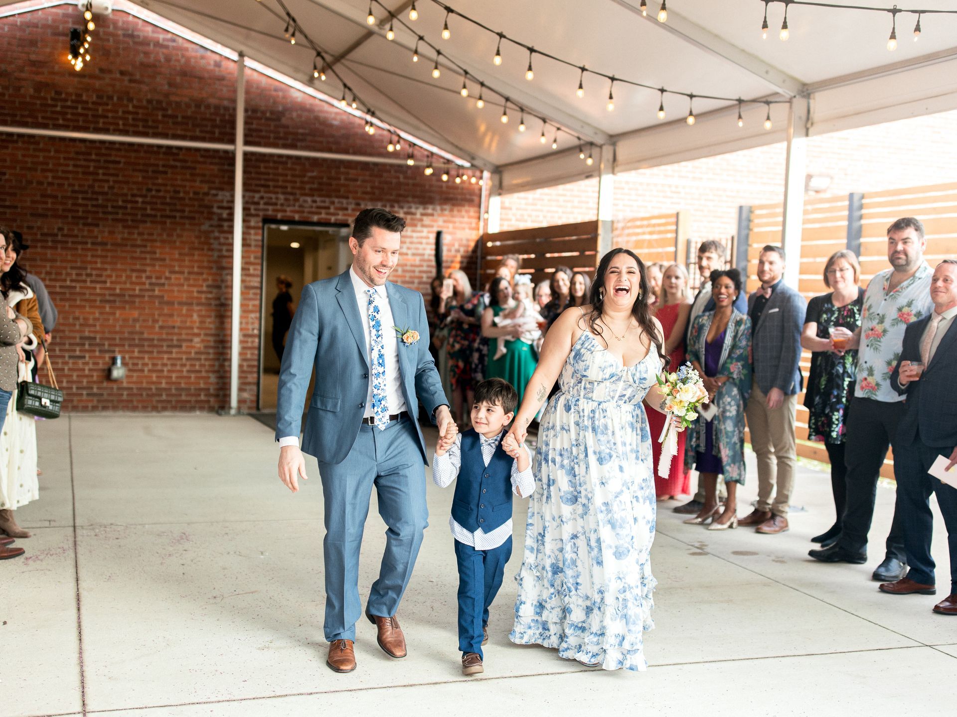 Newlyweds and child exit a wedding, smiling. Brick building and guests in background.