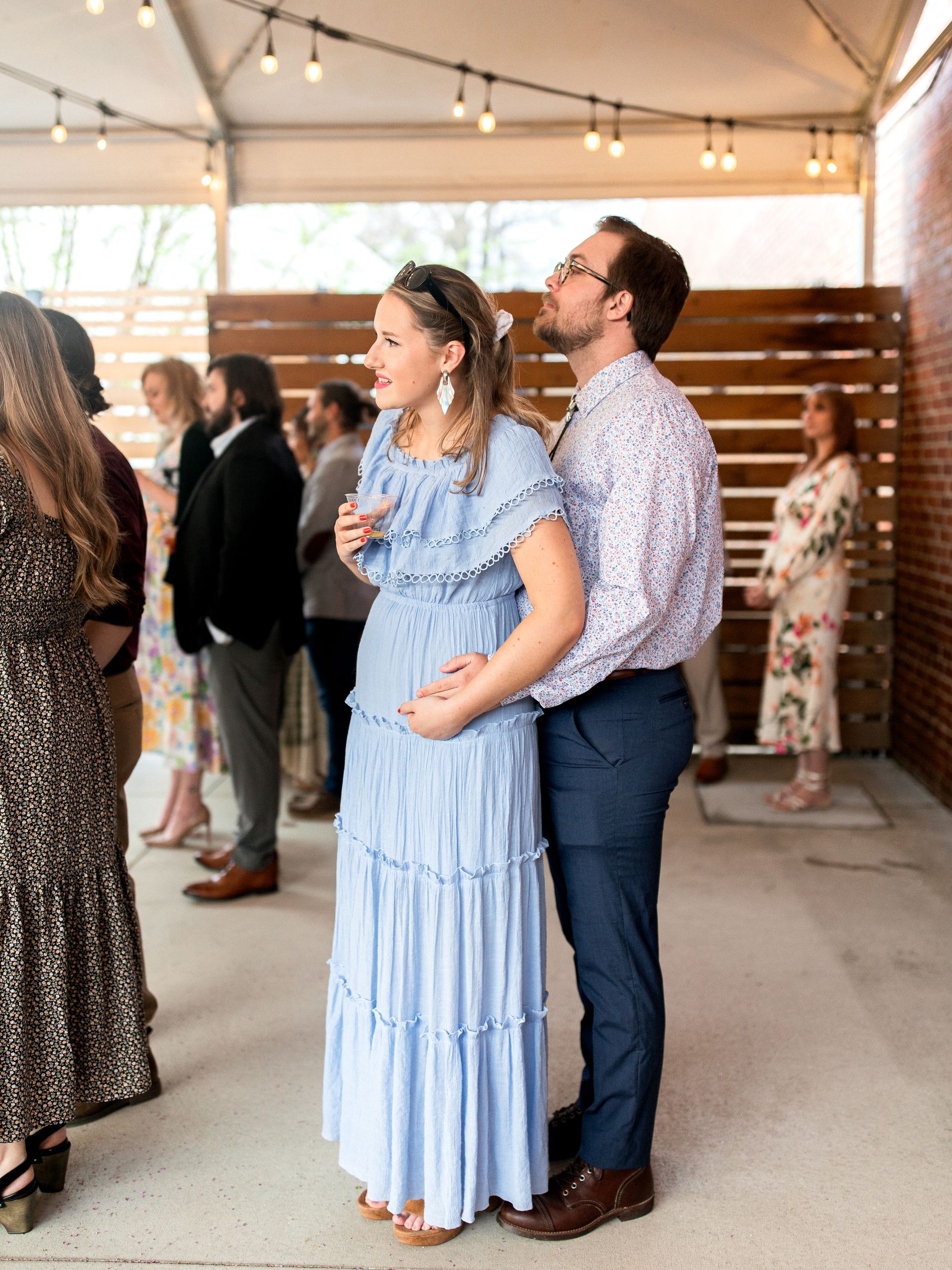 Pregnant woman in blue dress, embraced by man in floral shirt, at outdoor event.