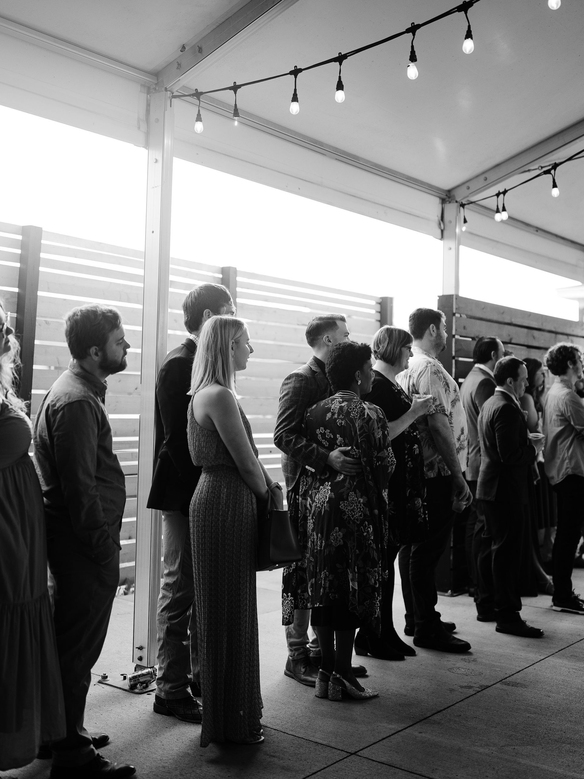 People standing, some in formal attire, under a covered patio with string lights; daytime.