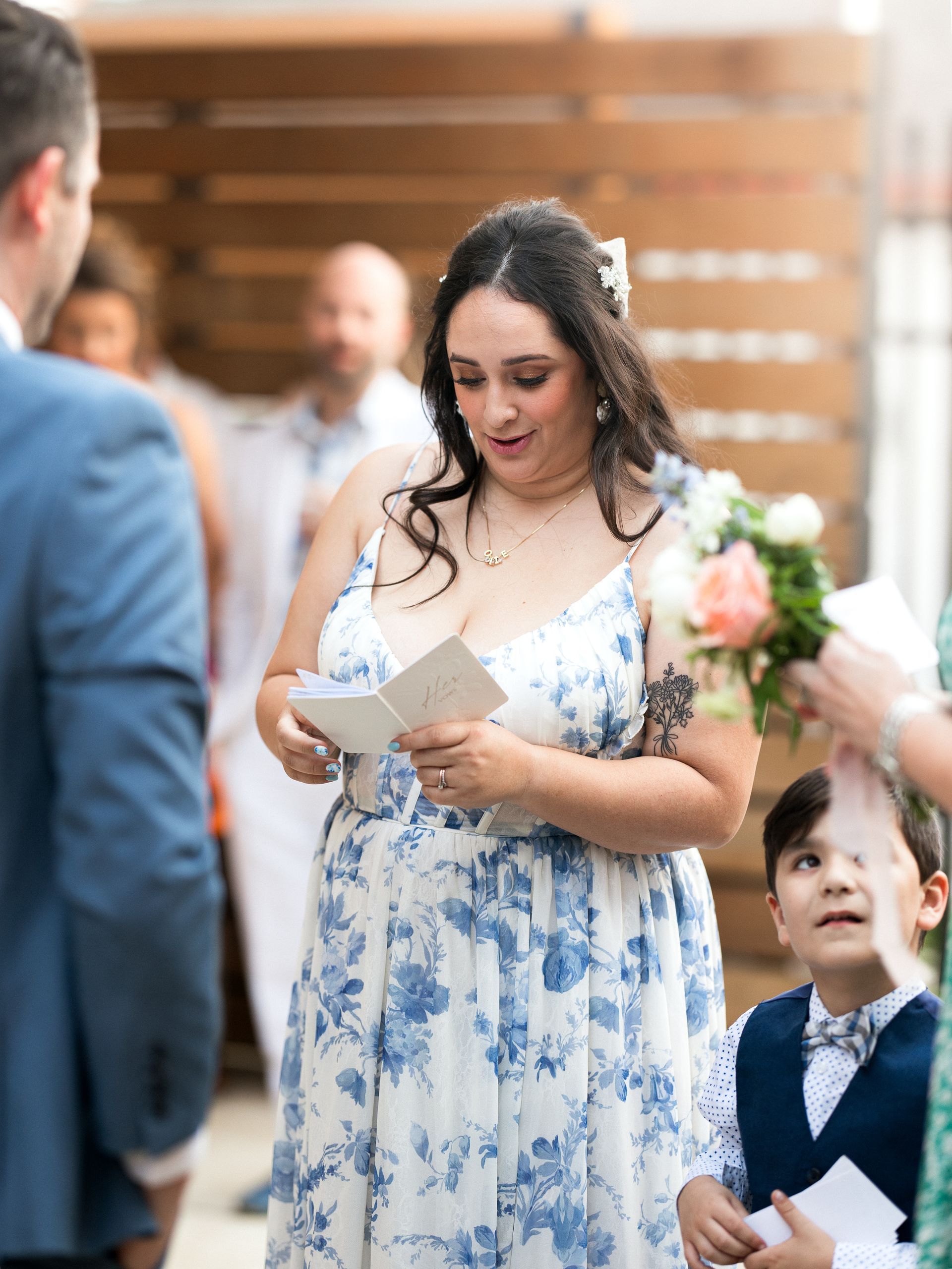 Woman in blue floral dress reads a card outdoors at a wedding; a boy looks up.