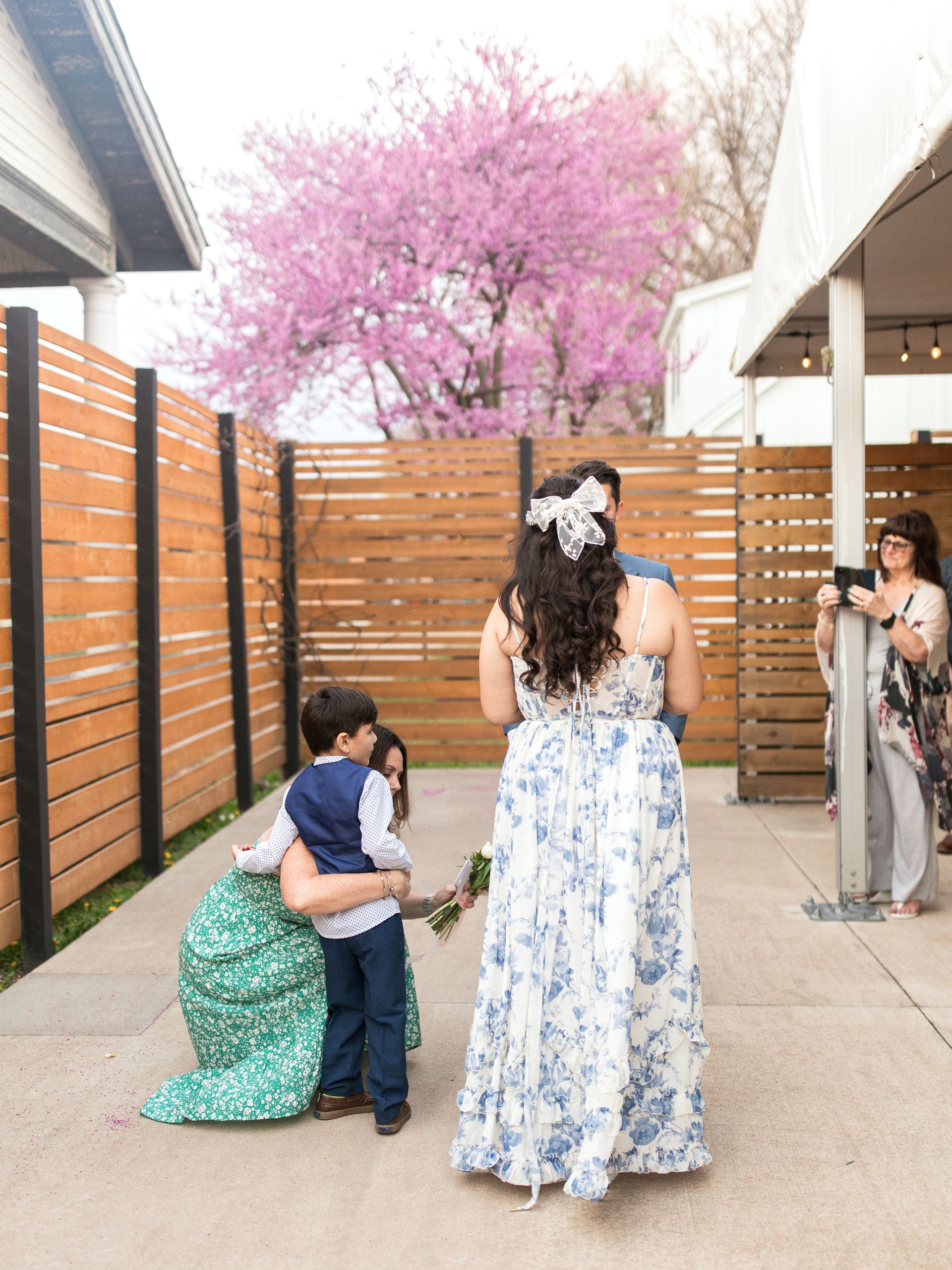Woman in a floral dress embracing a child, another person looks on, under a pink tree.