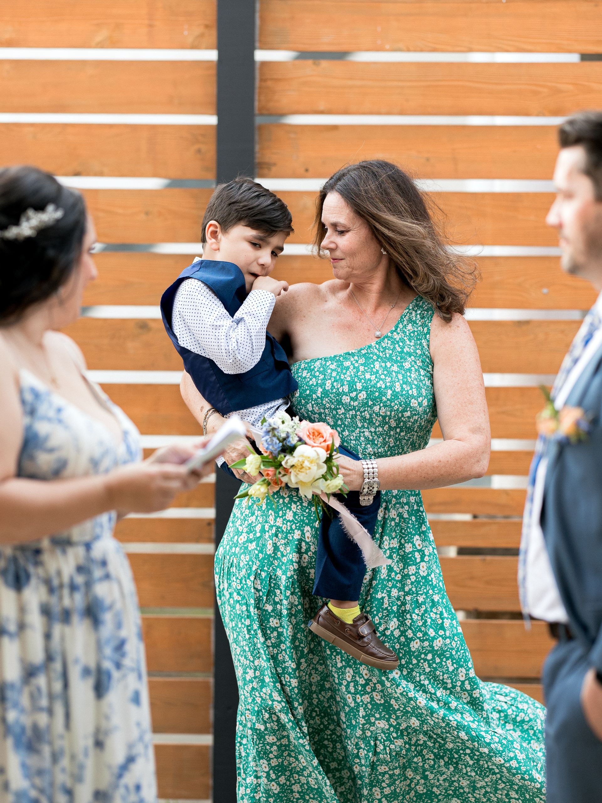 Woman in green dress holds a child during an outdoor ceremony, onlookers to the sides.
