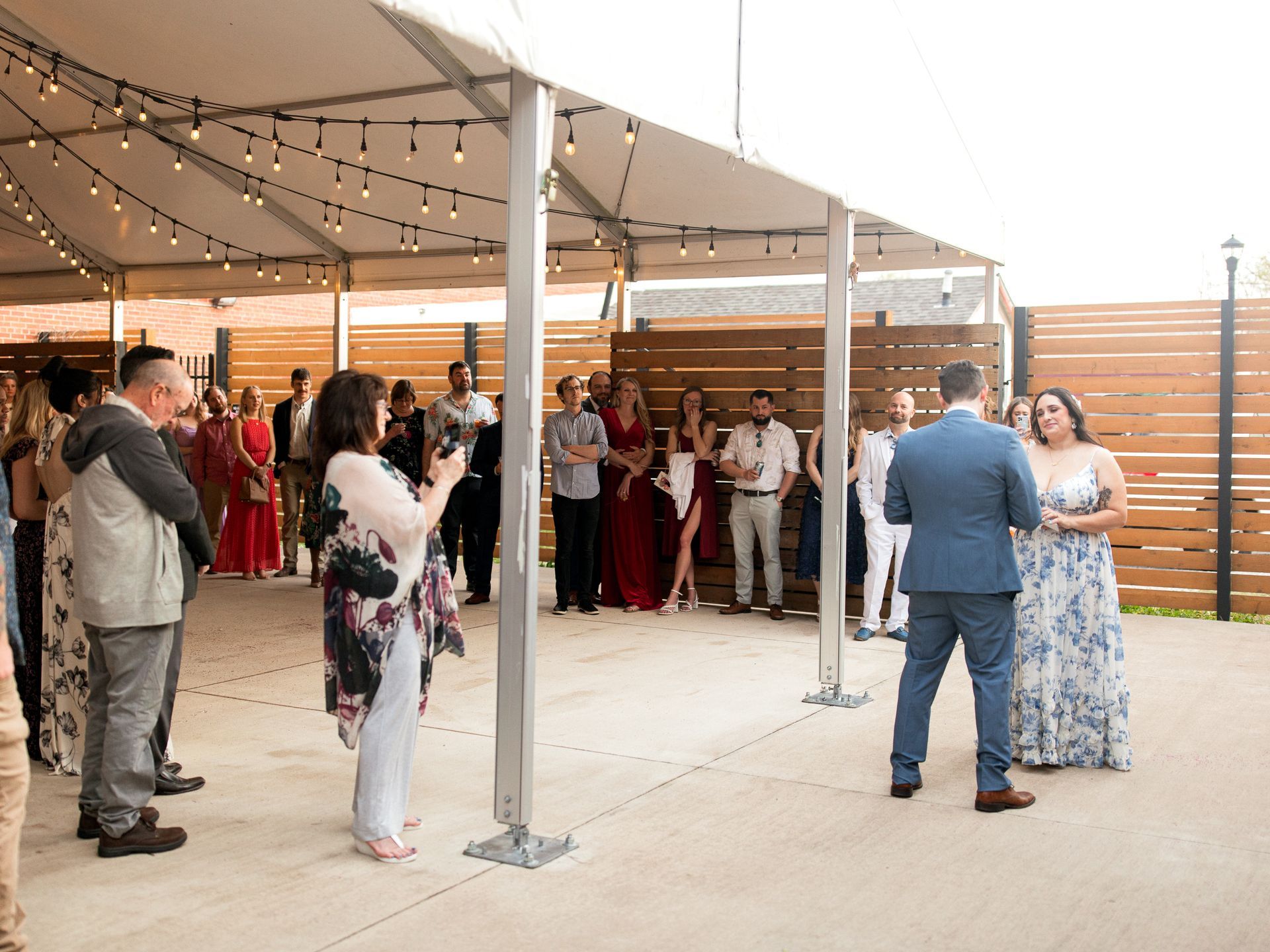 Couple stands at a wedding reception under a tent, surrounded by guests.