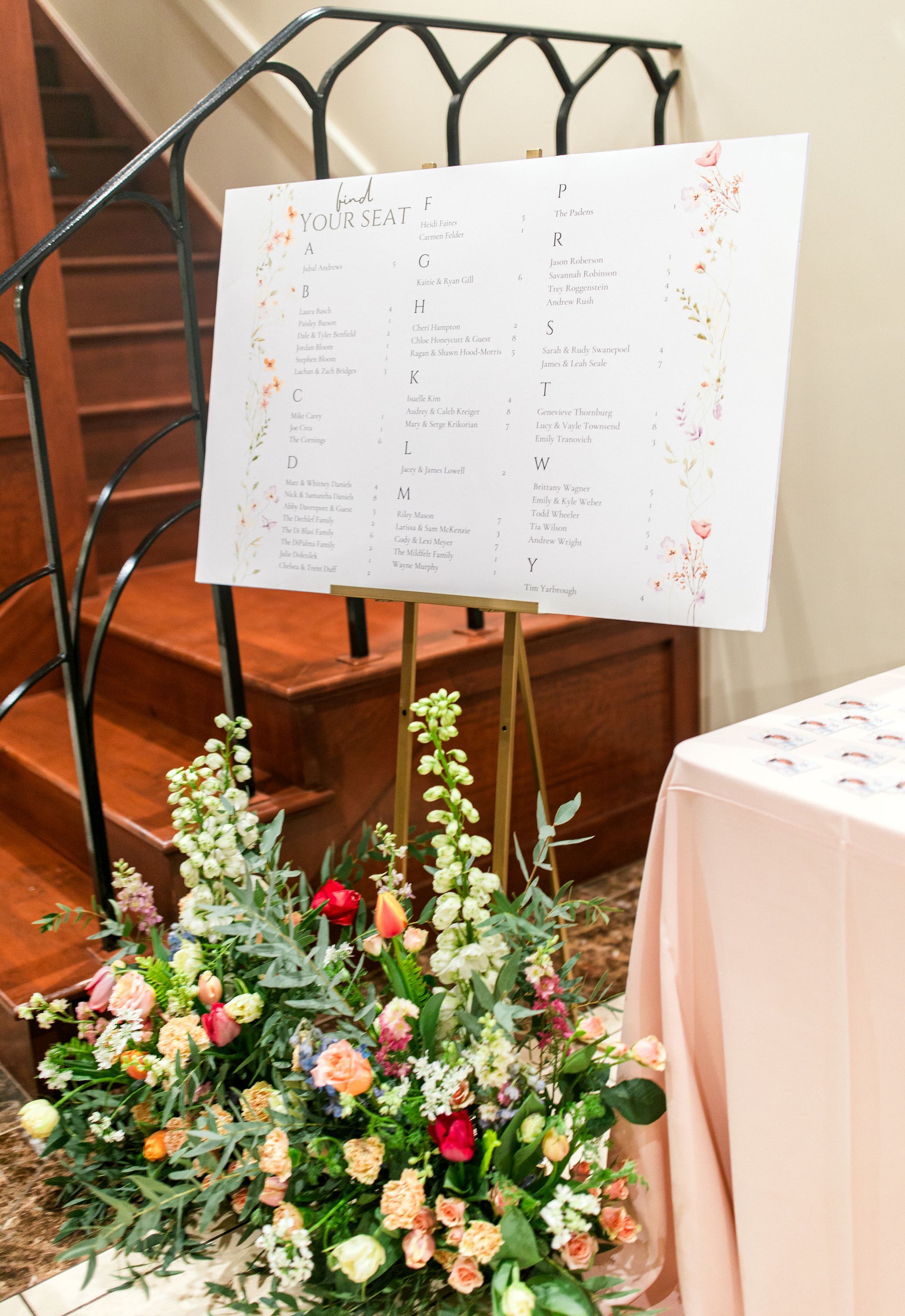 Seating chart on easel, surrounded by flowers at a wedding reception. Stairs in the background.