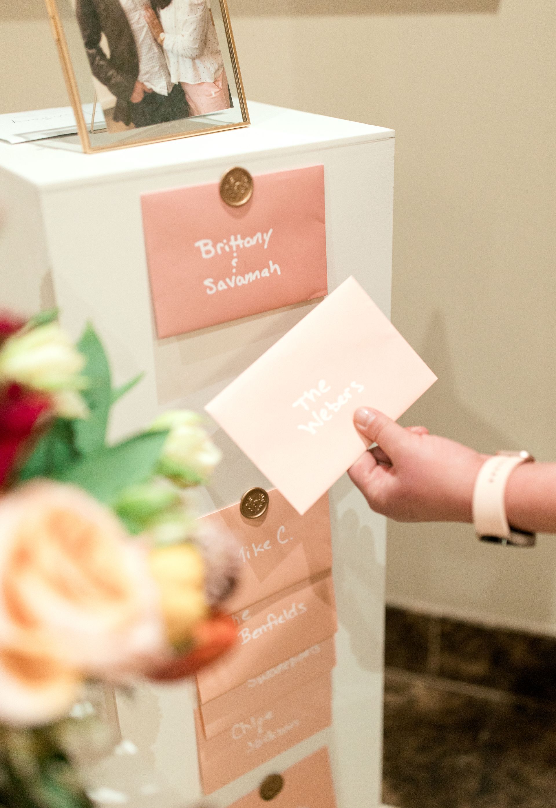Person holding a pink envelope, next to a display of envelopes with names, near a flower arrangement and framed photo.