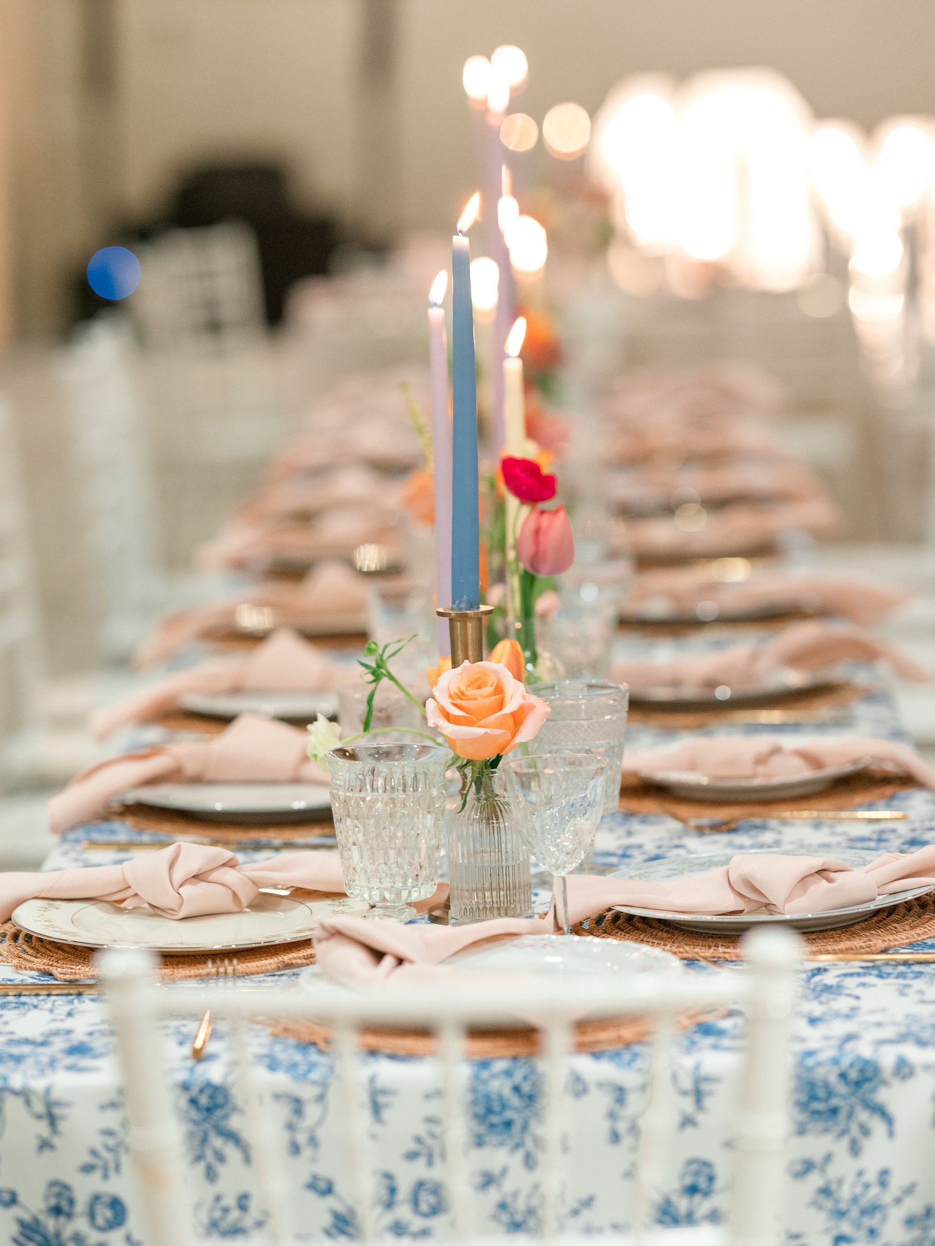 Elegant table setting with candles, flowers, and pink accents on blue floral tablecloth.