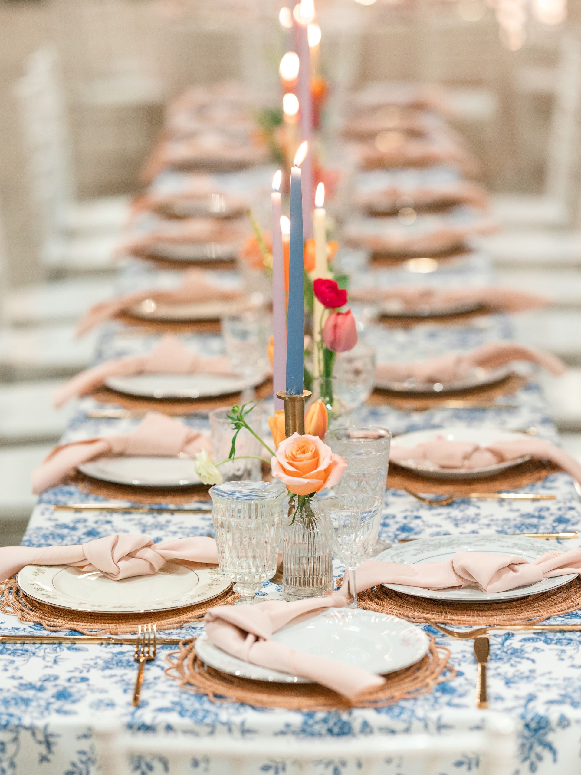 Long table setting with floral tablecloth, pink napkins, gold accents, and colorful candles.