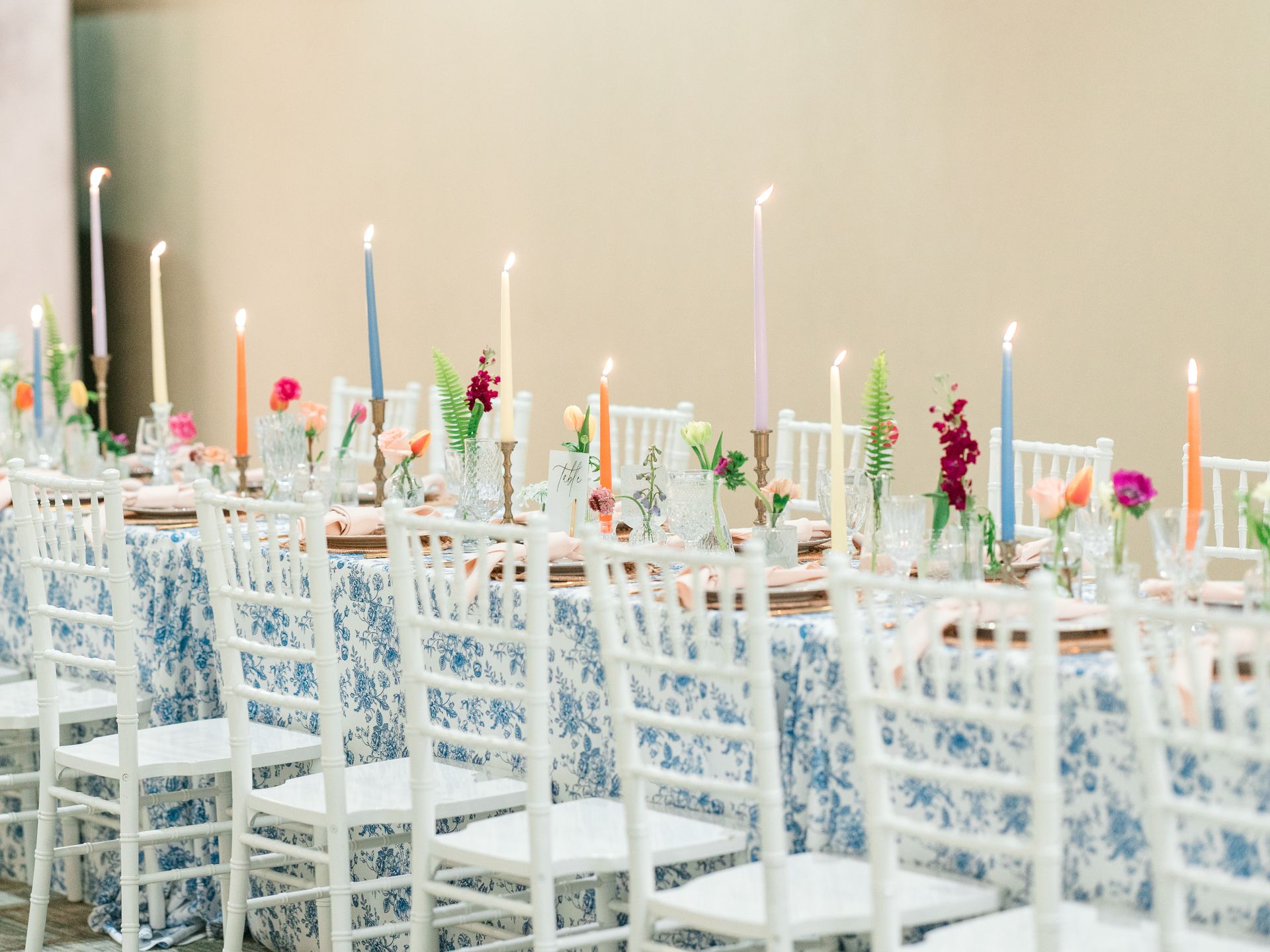 Long table set for a celebration with blue floral tablecloth, white chairs, colorful candles, and floral centerpieces.