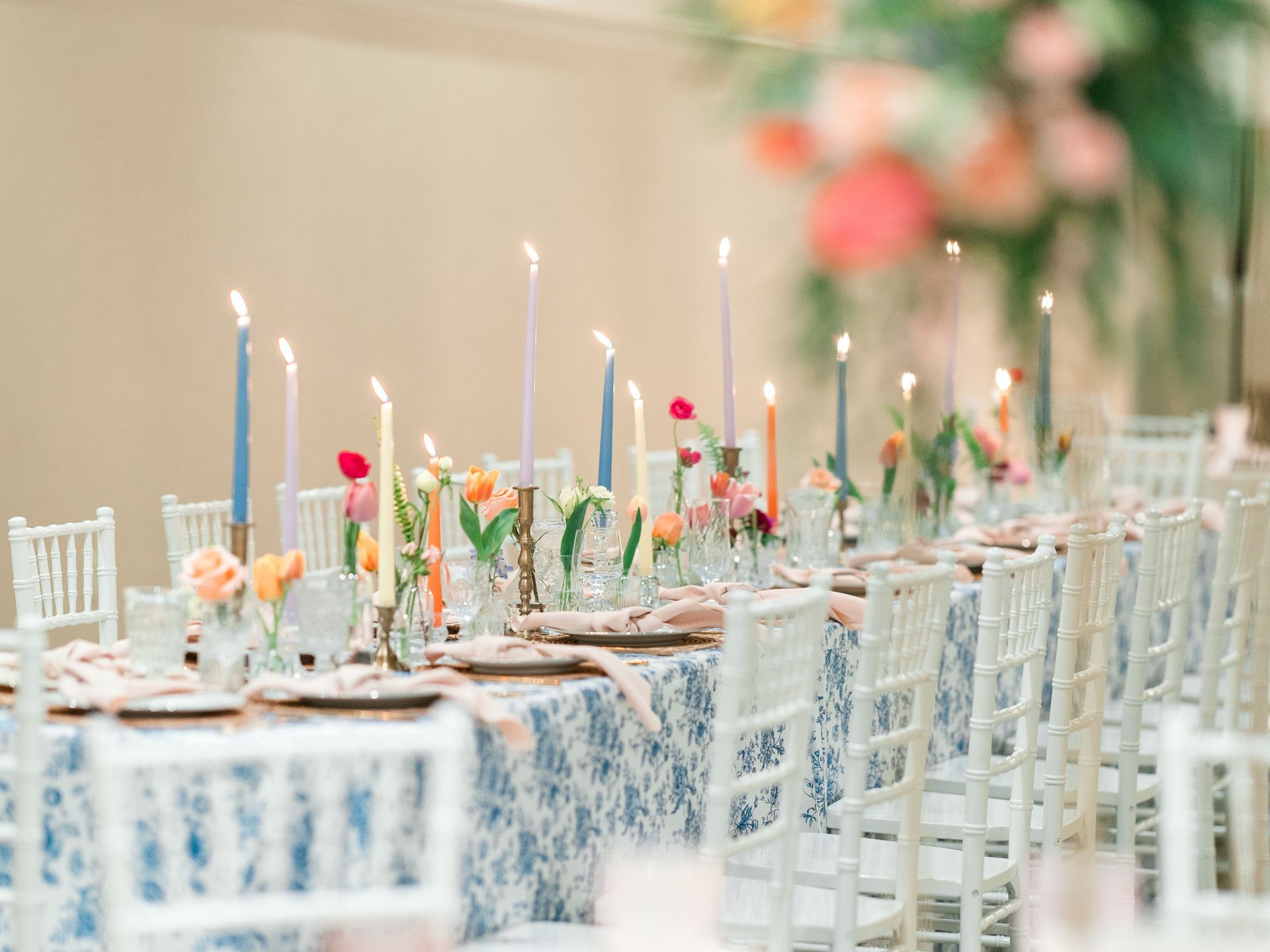Long table set for an event with colorful candles, floral arrangements, and white chairs.