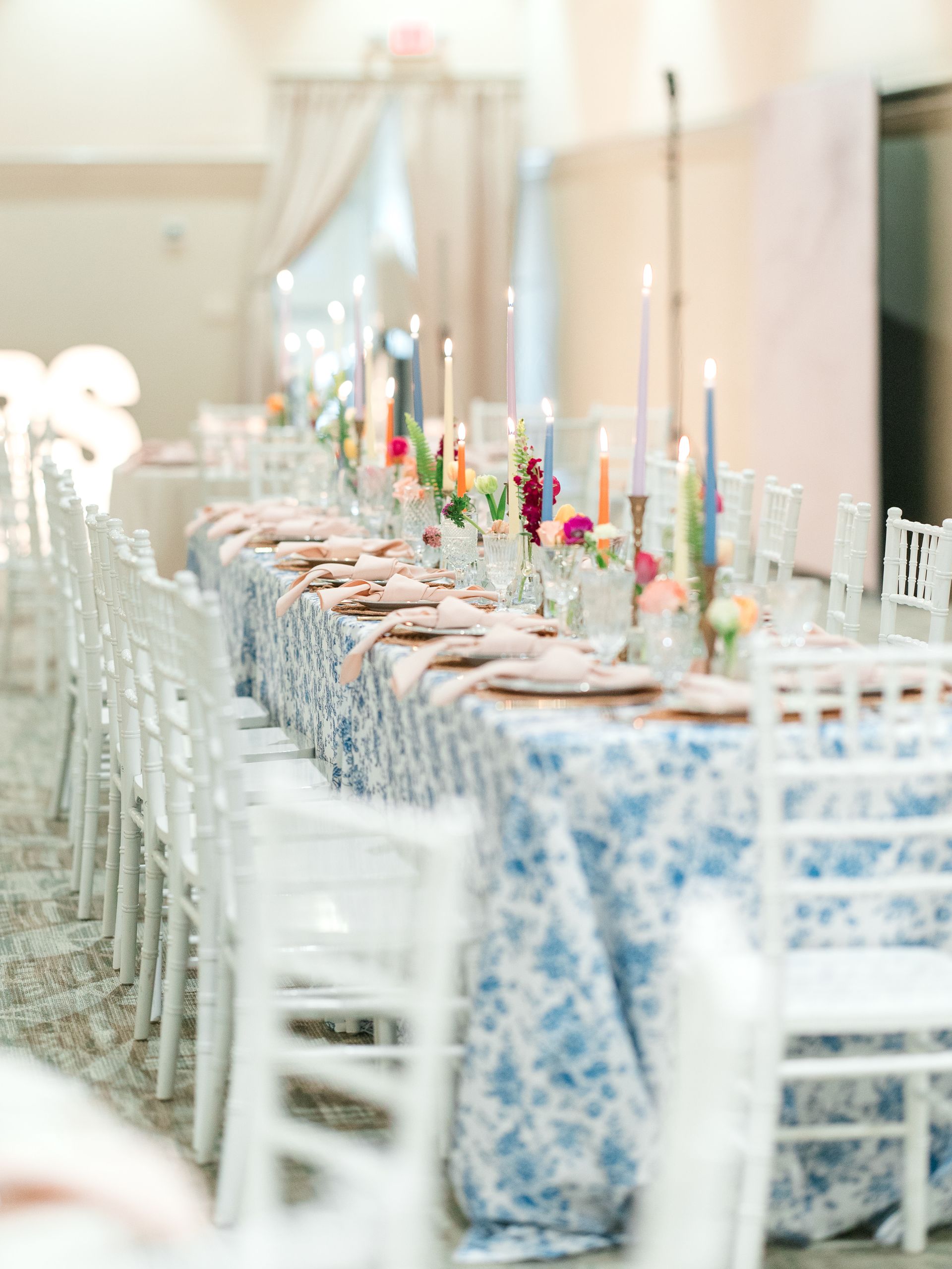 Long table setting with blue floral tablecloth, colorful candles, and white chairs.