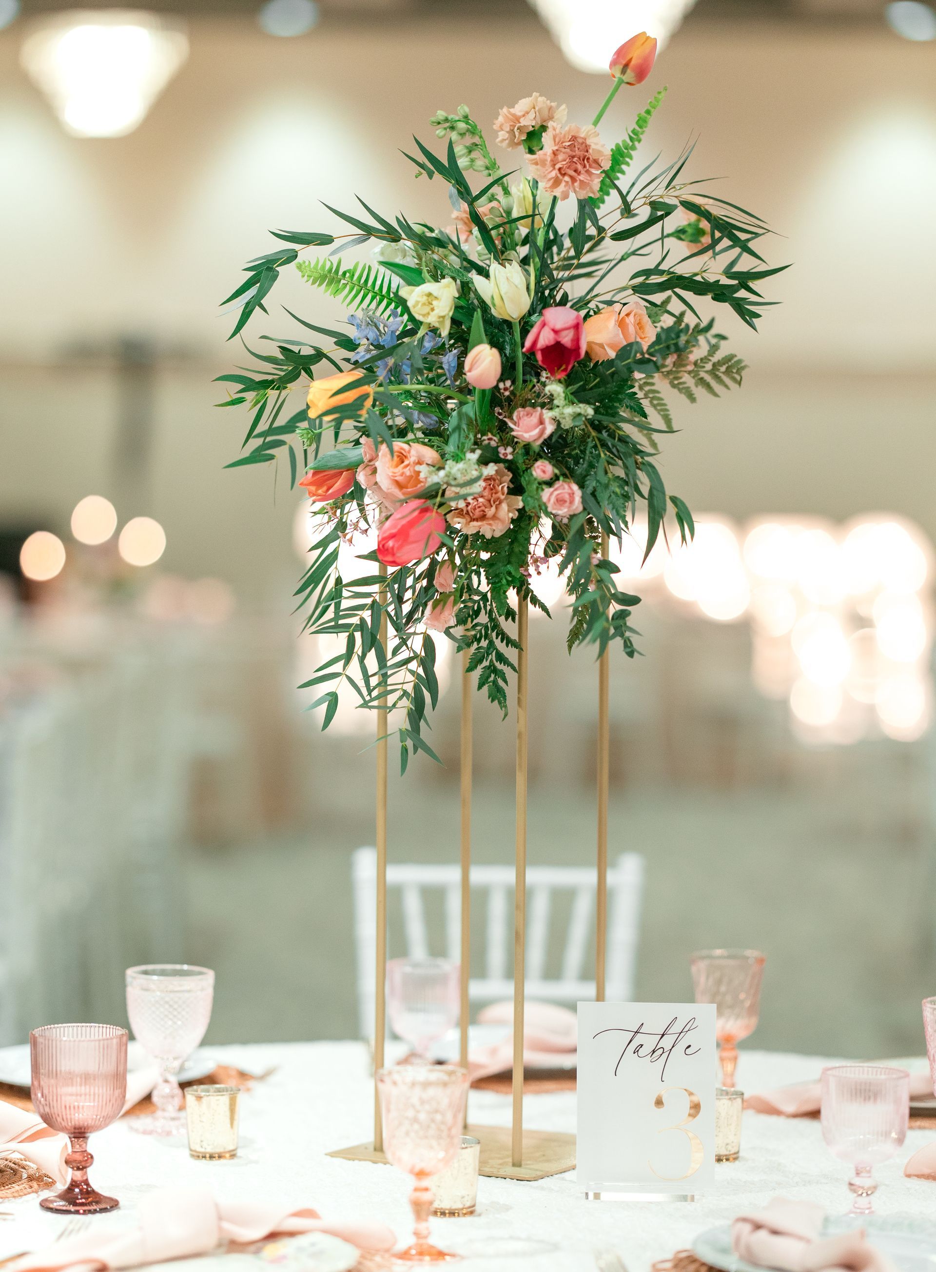 Tall floral arrangement on a table with place settings, gold stand, and blush pink accents.