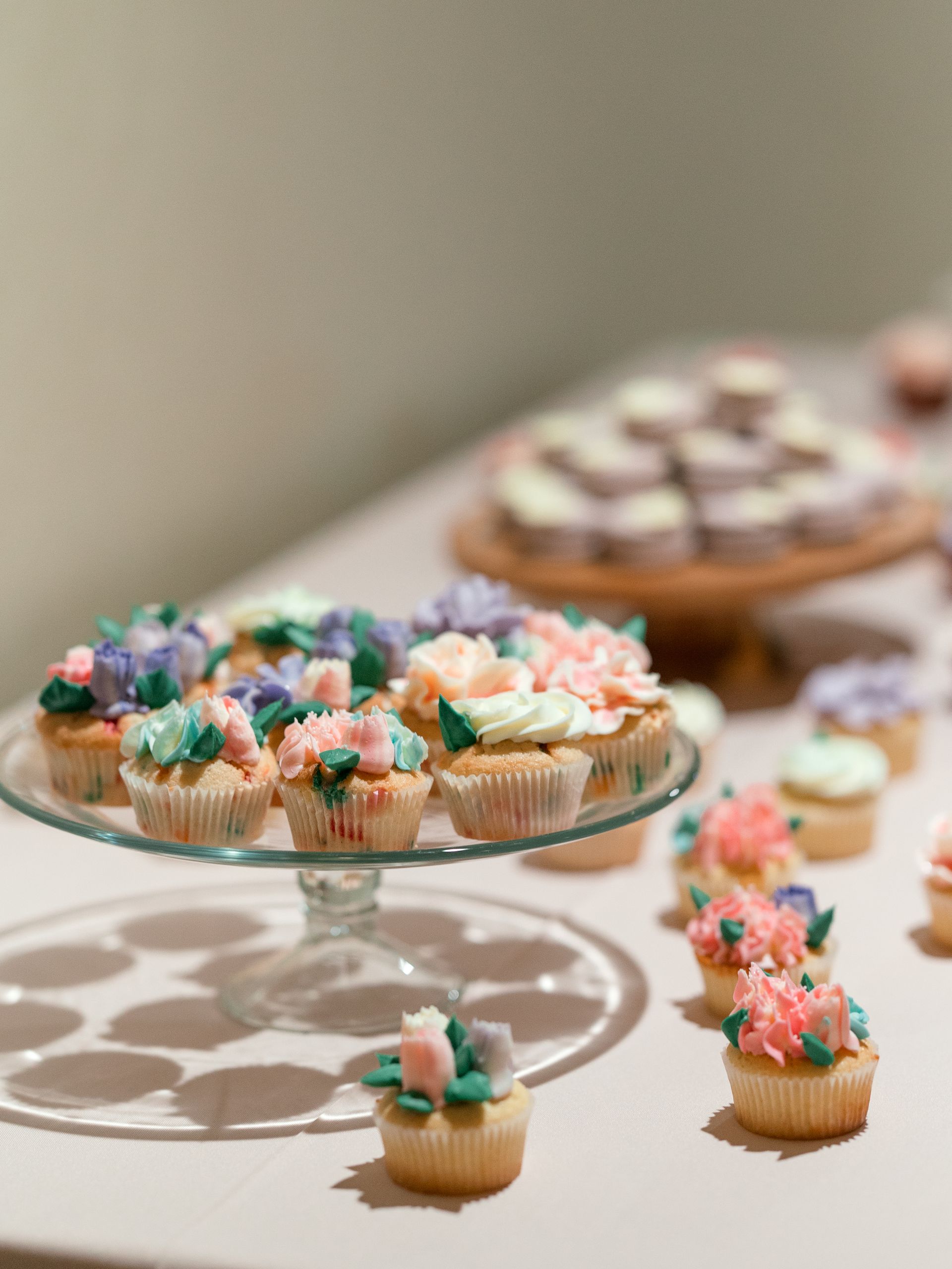 Cupcakes with floral frosting on a glass stand, and more cupcakes and treats on a table.