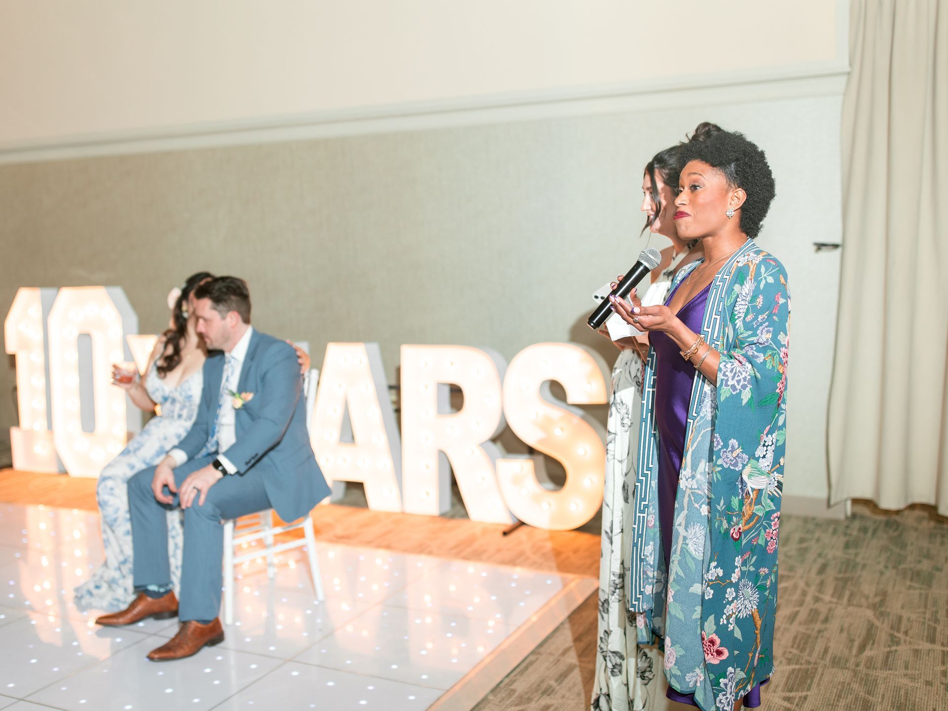 Woman with microphone speaks near seated couple in front of lit-up 