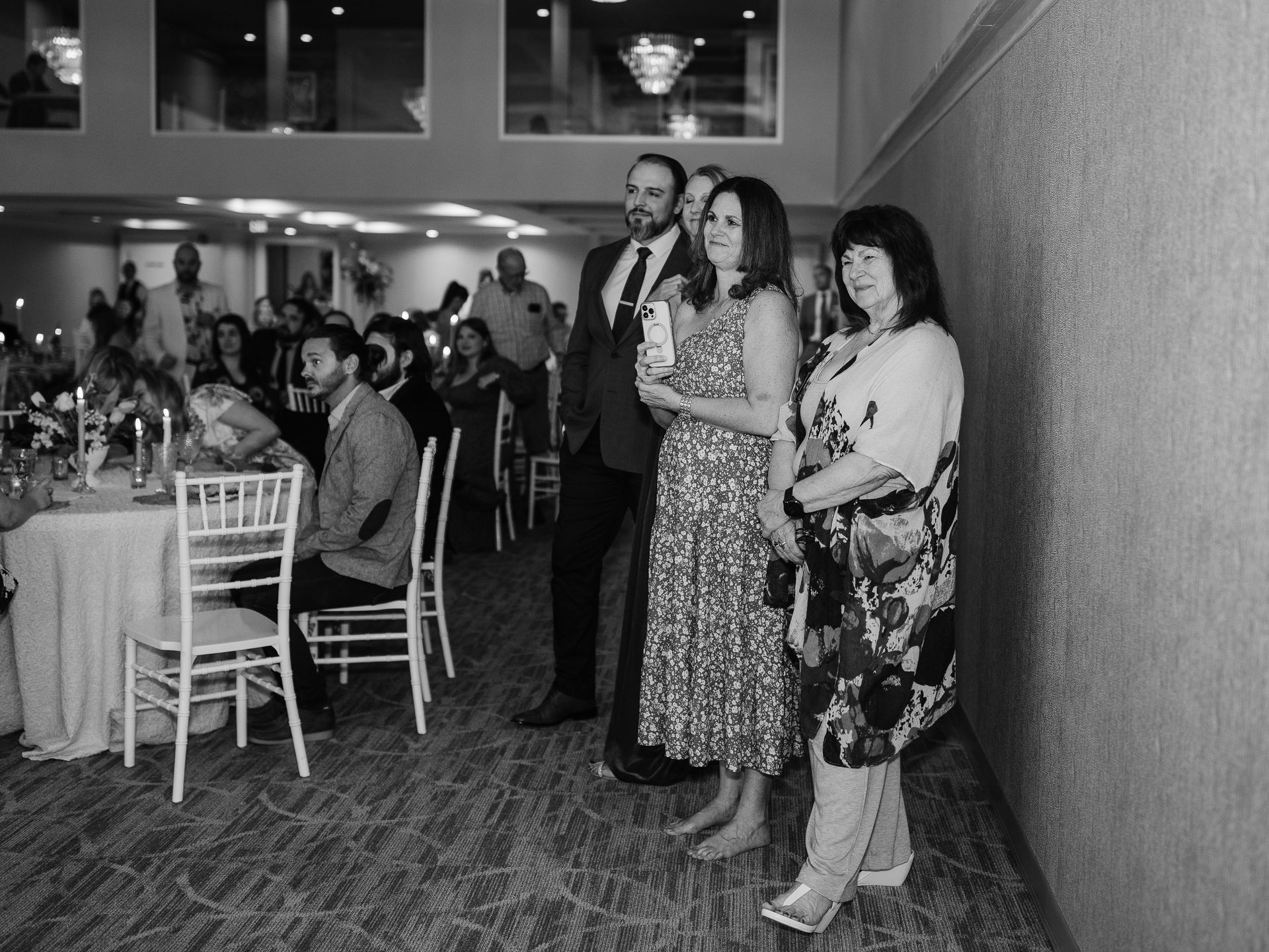 People standing in a room with a party. Two women and a man are in the foreground, near a wall.