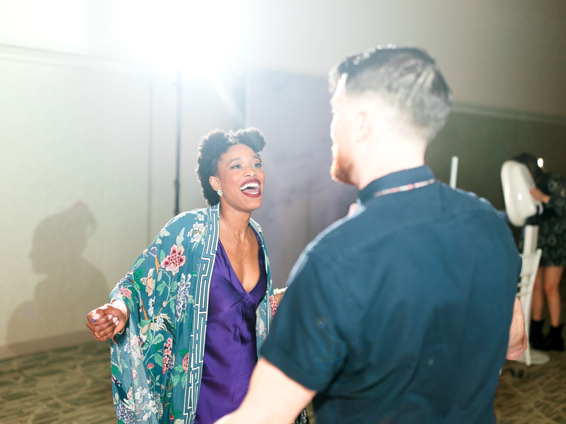 Woman in floral robe laughs, facing a man in a blue shirt. Lit room, blurred figures in the background.