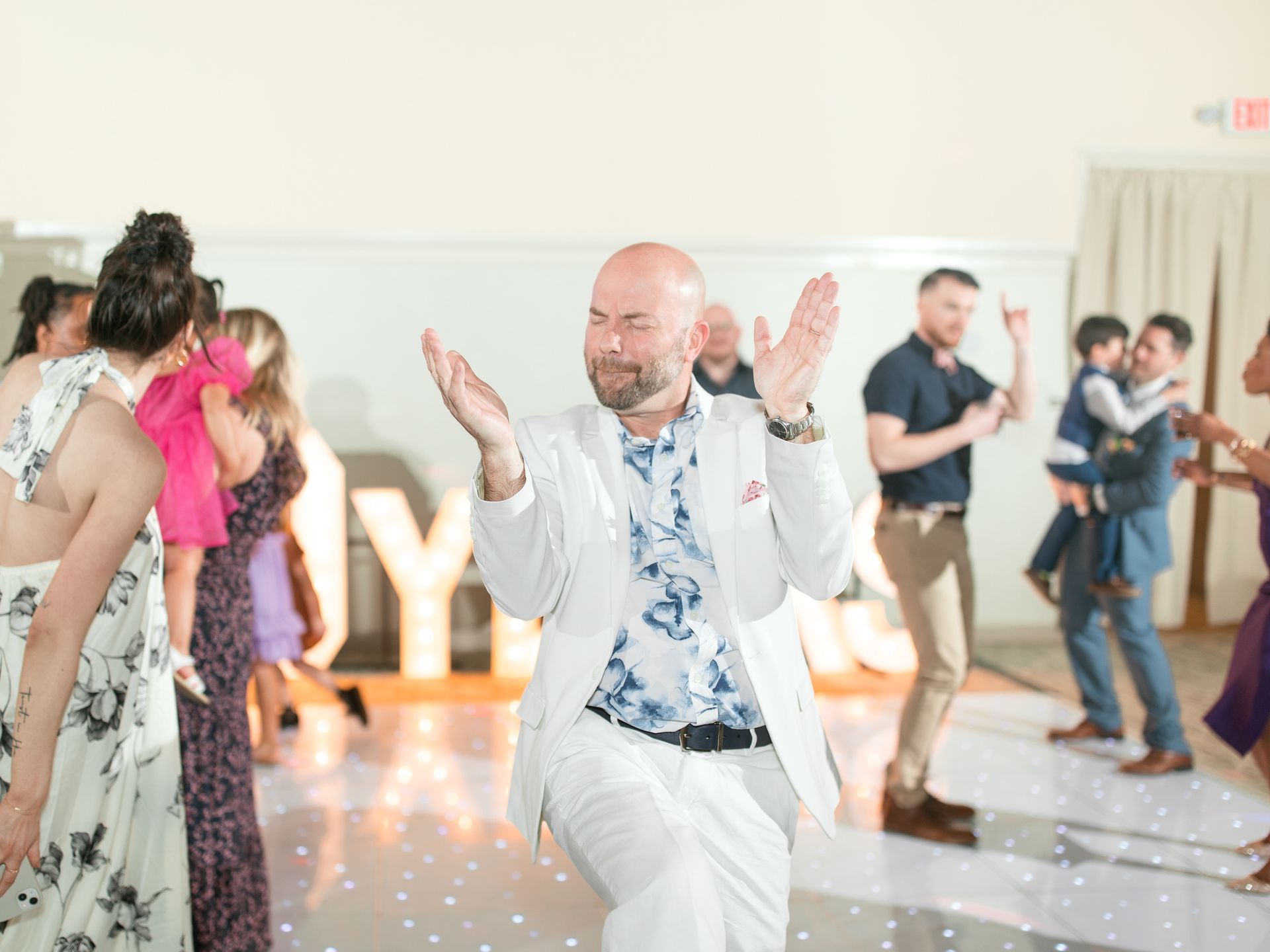 Man in white suit dances on a lit dance floor, arms raised. People in the background.