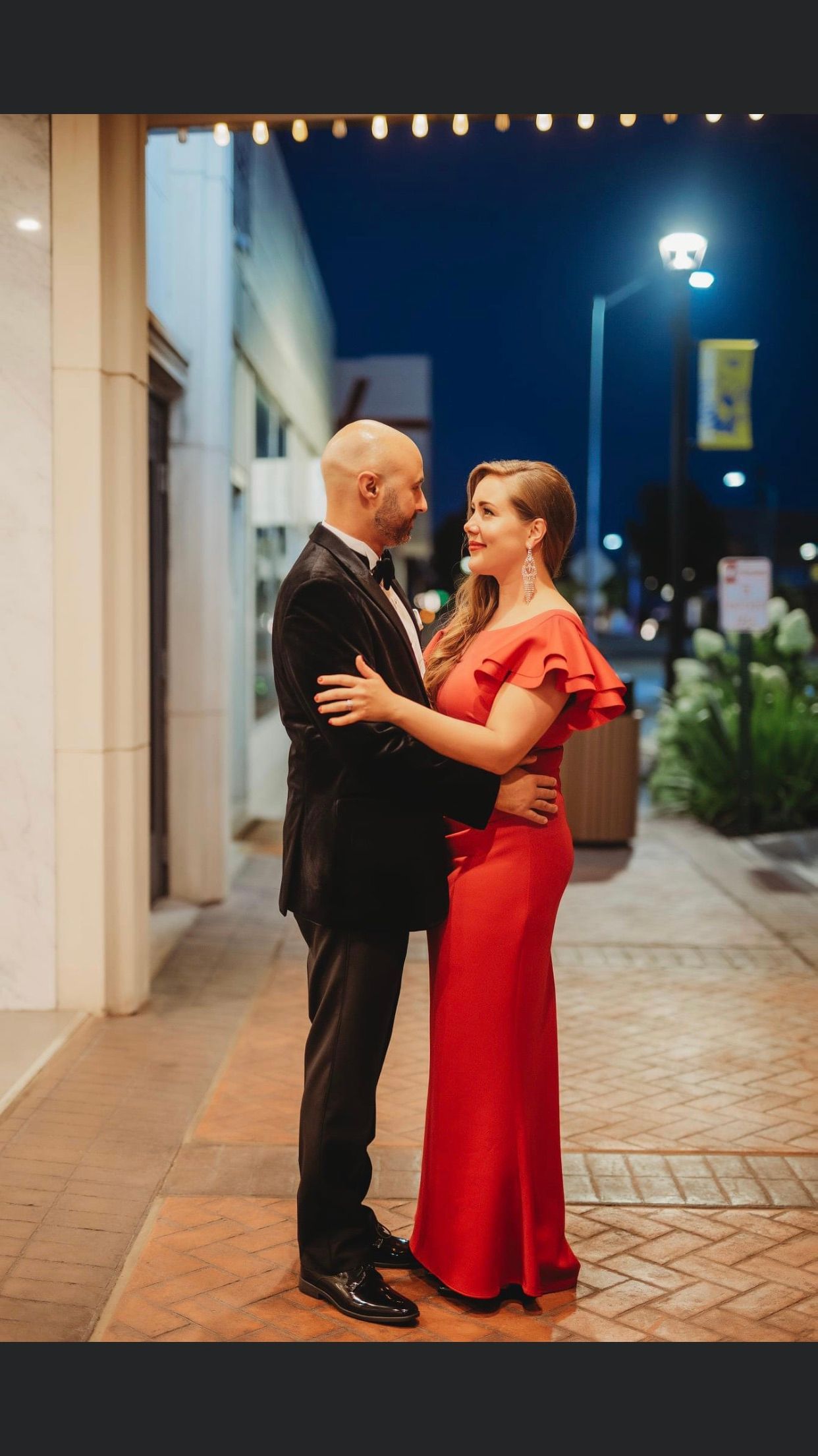 Couple in formal attire embracing, looking at each other outdoors at night. Man in a tuxedo, woman in a red gown.