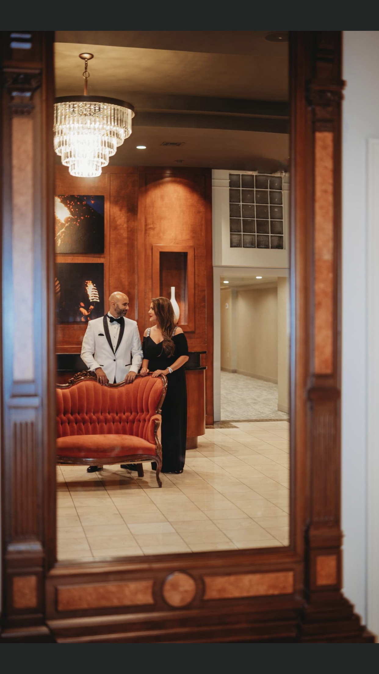 Couple in formal attire reflected in a large ornate mirror, standing near a red couch in a hallway.