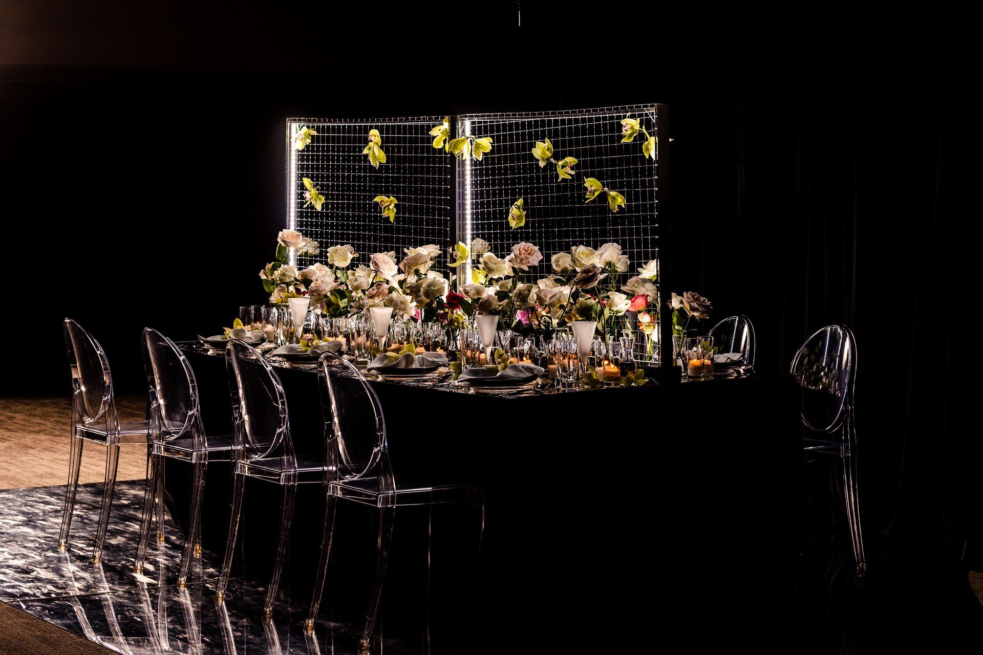 Elegant black dining table set with clear chairs and floral centerpiece against a modern grid backdrop.