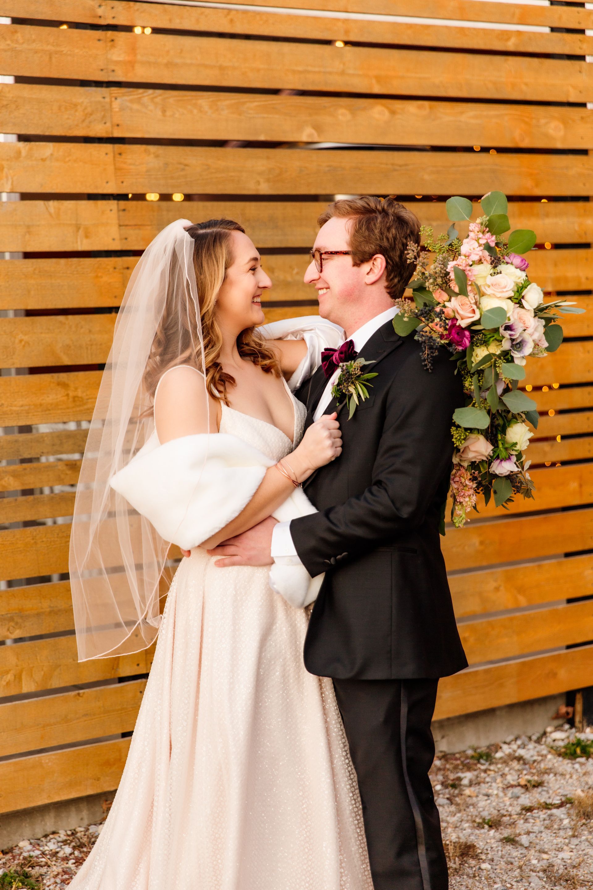 Bride and groom embracing, smiling, against a wooden fence. She wears a veil and shawl, he a tuxedo with a floral bouquet.