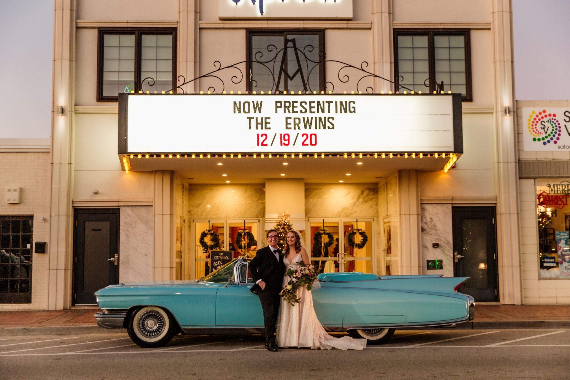 Couple poses by a light blue vintage car in front of a theater sign that reads 
