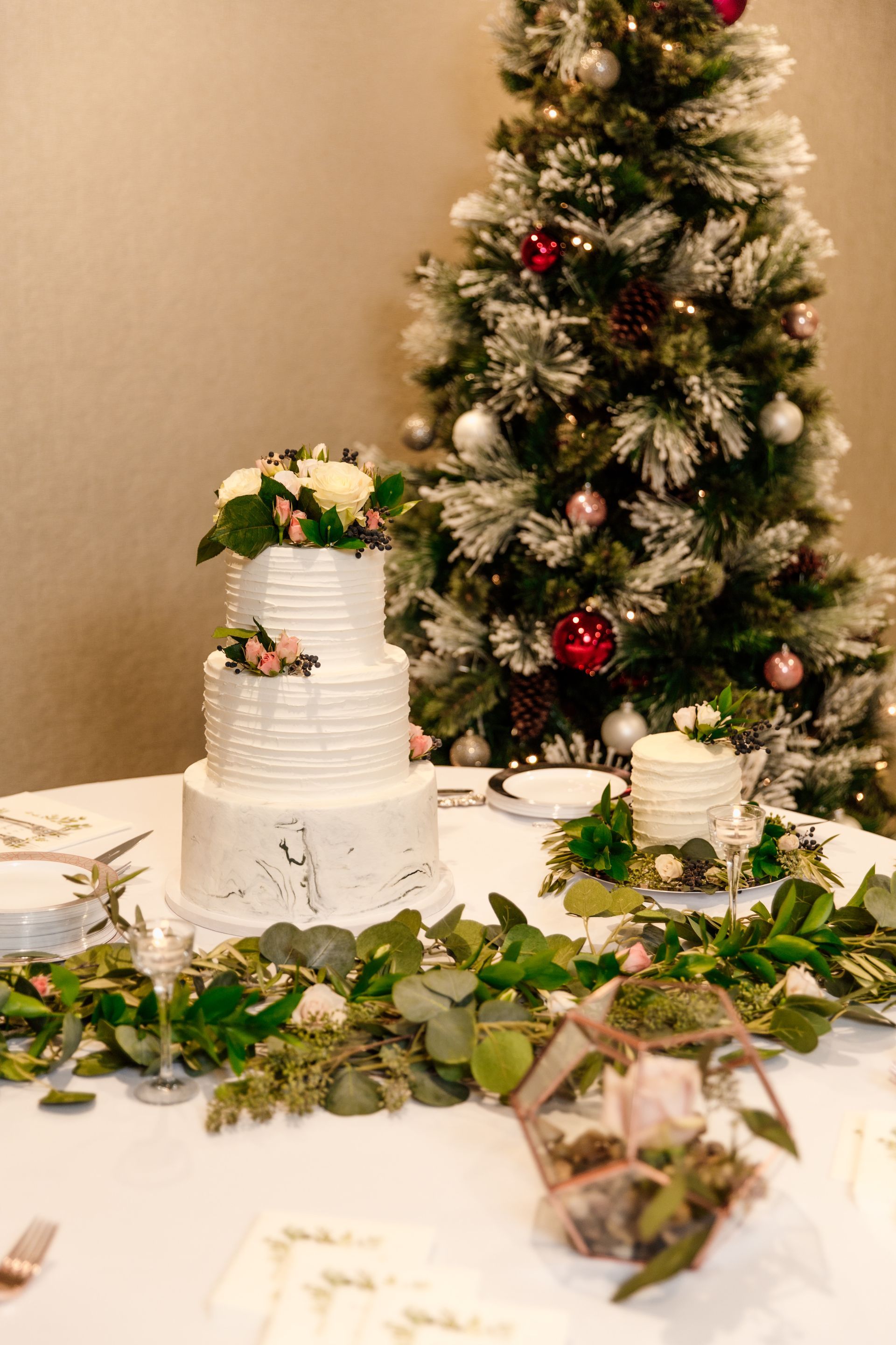 Three-tiered white wedding cake with floral topper on a table decorated for Christmas, with tree in background.