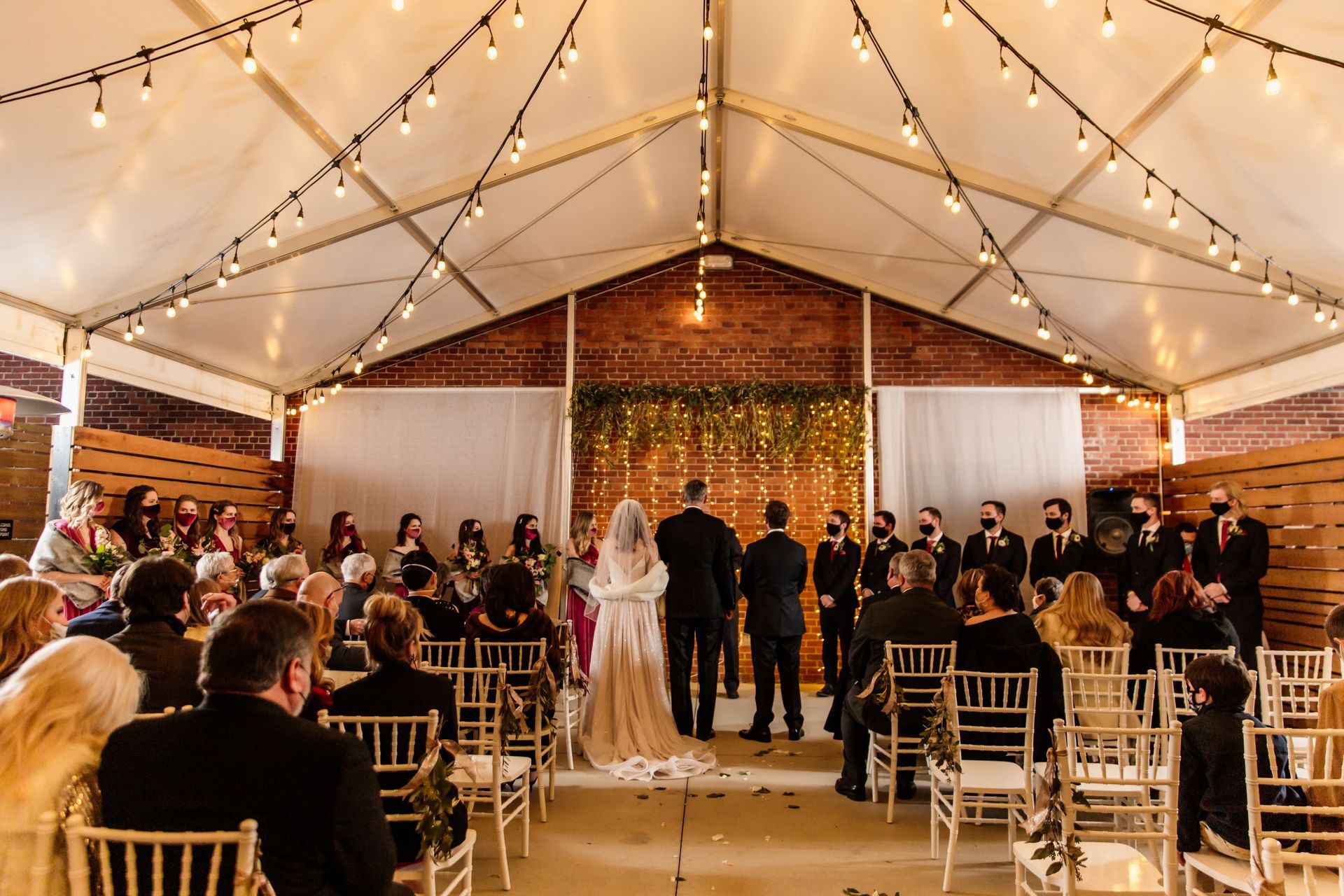 Women in a wood-paneled room, conversing and holding drinks near a floral arrangement, with a window and bar area in view.